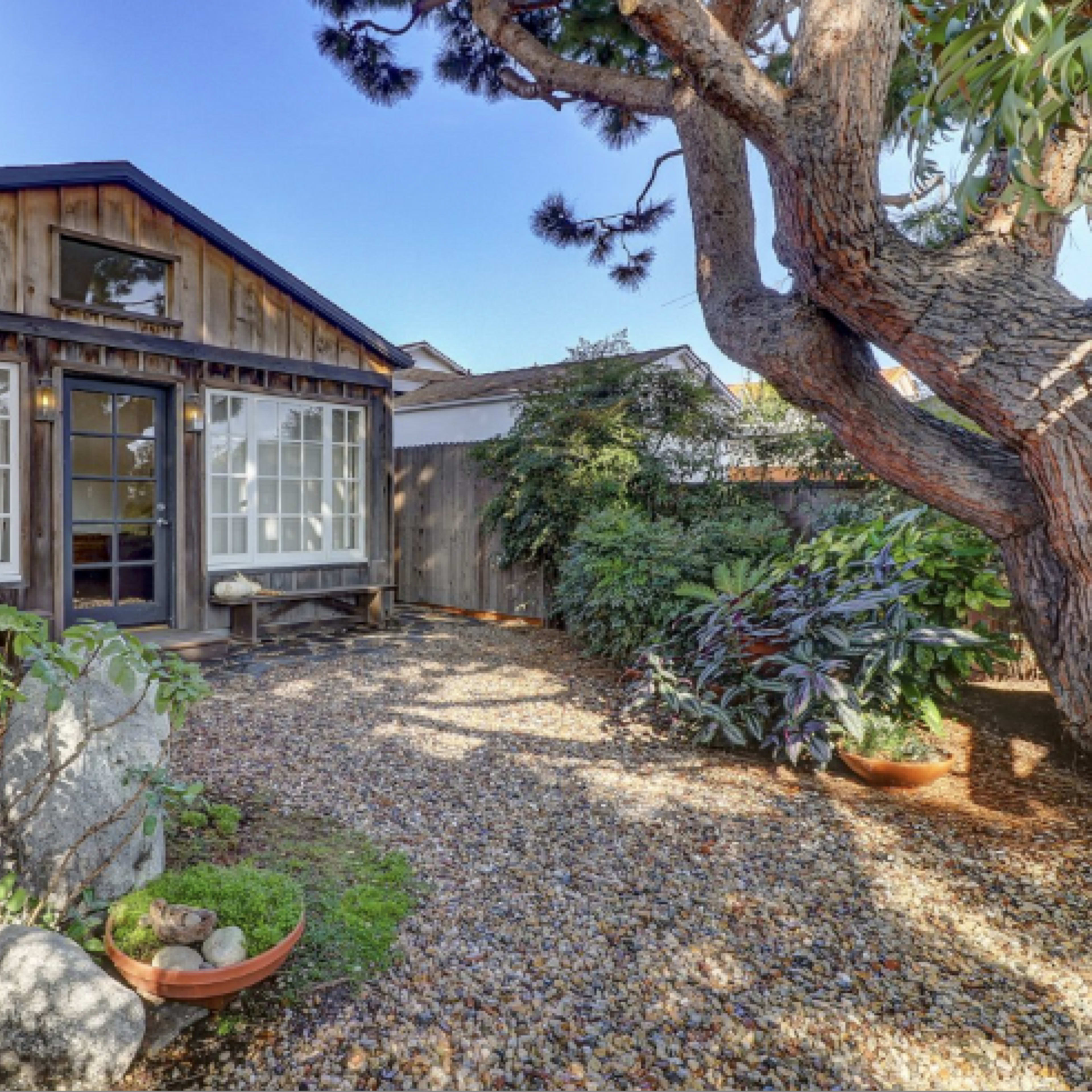 A wooden cabin with large windows is surrounded by a landscaped garden featuring gravel pathways and various plants.