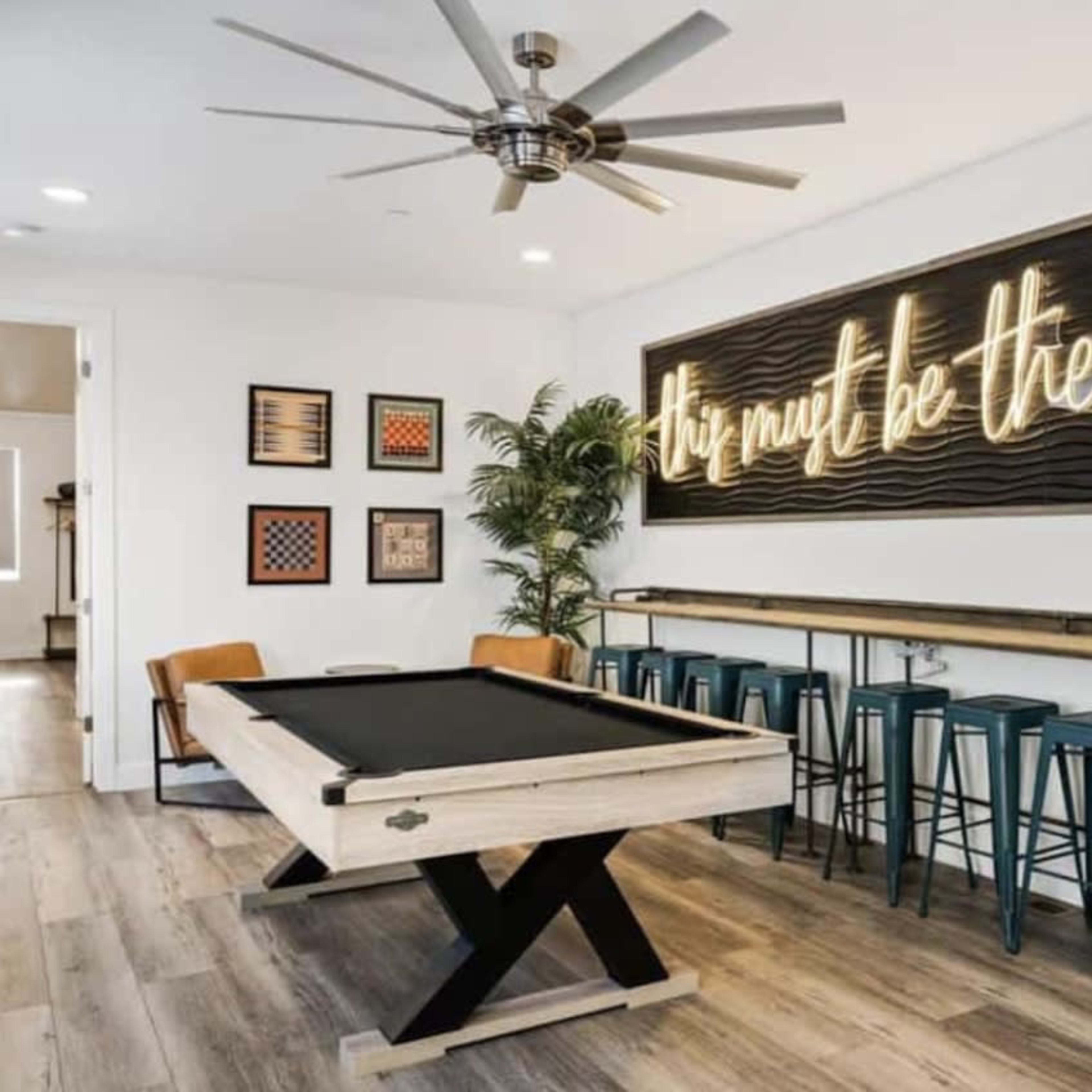 A game room with a pool table, a neon sign on the wall, and seating at a bar-style table.