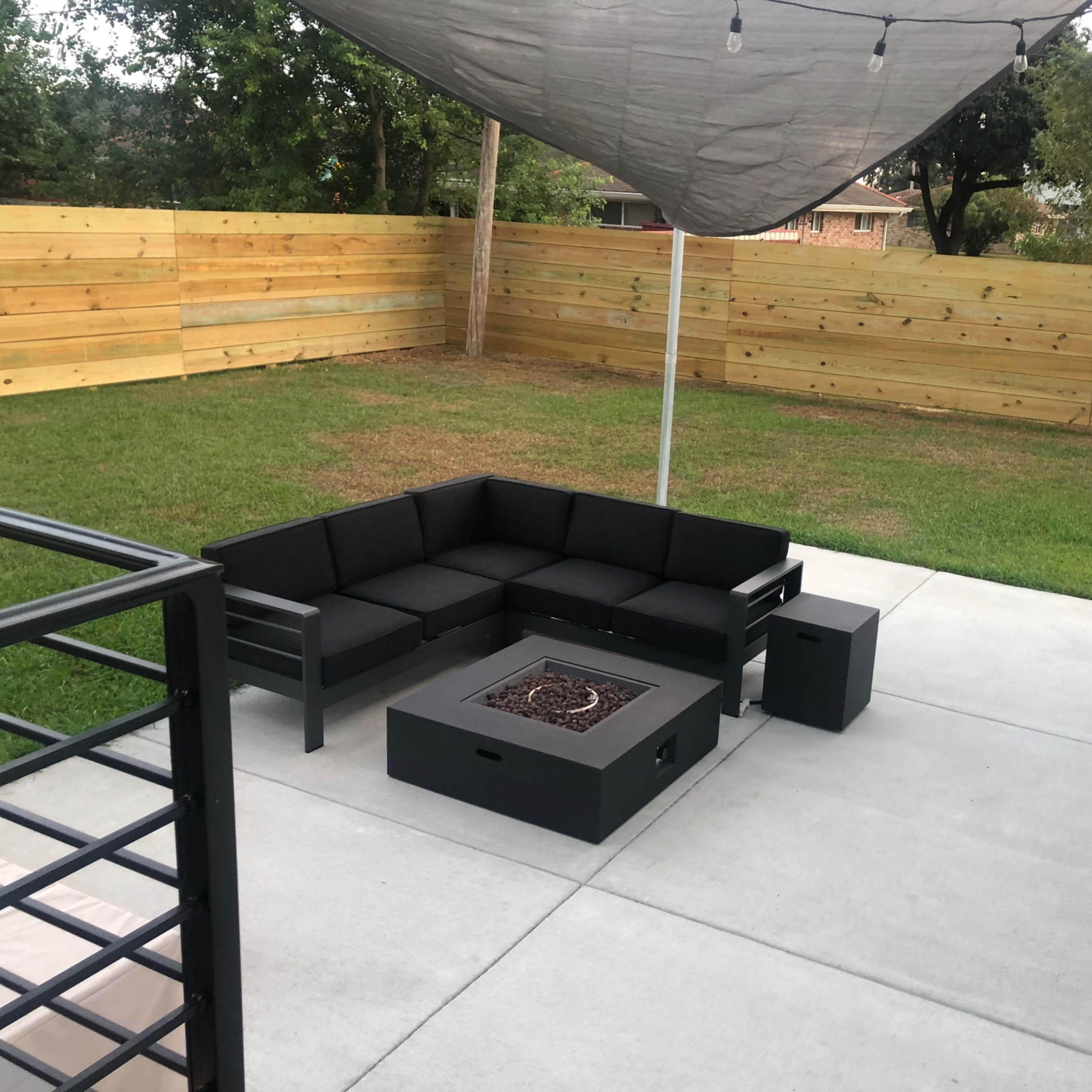 A modern outdoor seating area features a black sectional sofa and a fire pit under a shaded canopy on a concrete patio, surrounded by a wooden fence and grass.