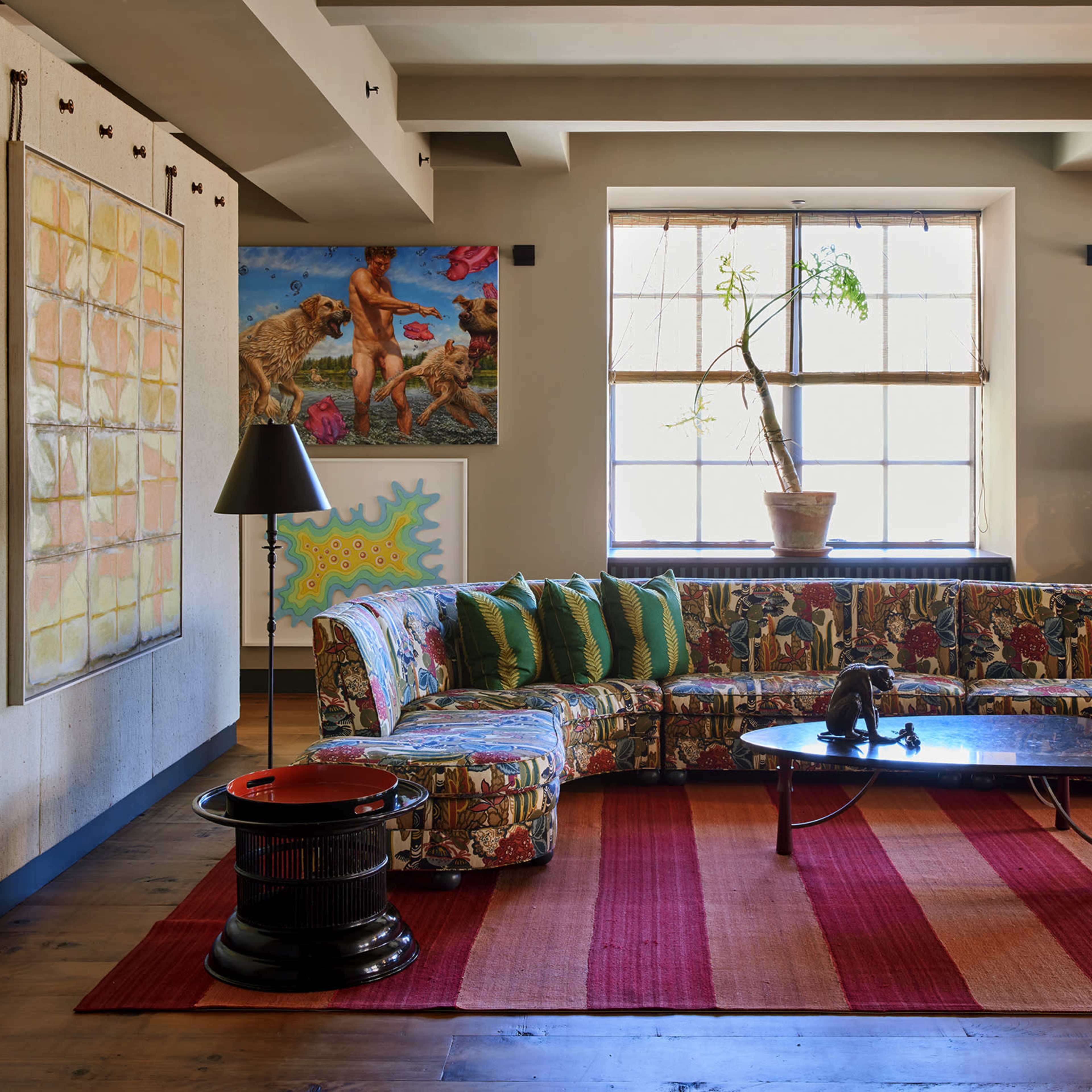 The room features a colorful patterned sectional sofa, a round coffee table, and various pieces of artwork on the walls, with a large window providing natural light.