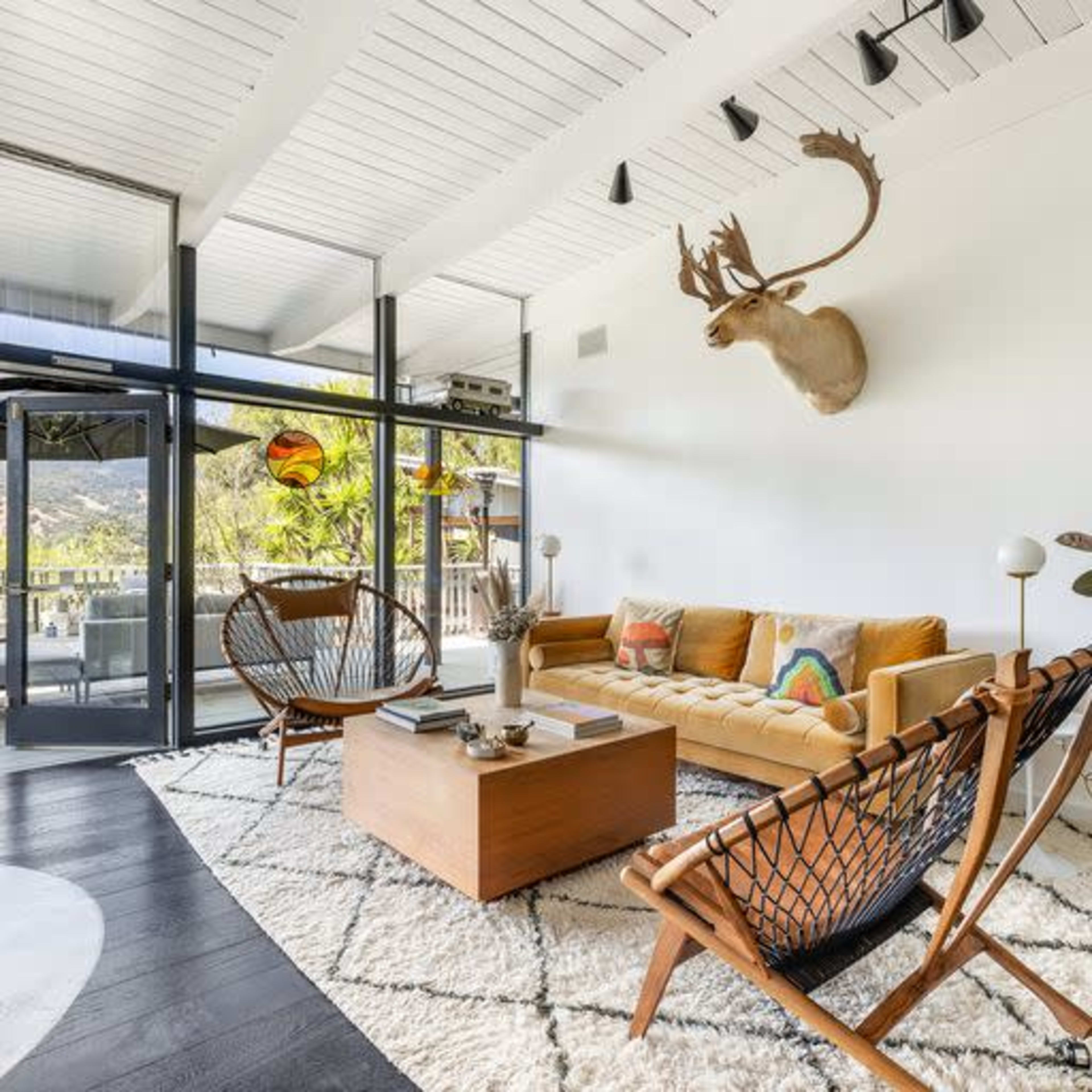 A modern living room featuring a yellow sofa, two woven chairs, a coffee table, and a mounted deer head on the wall, with large windows allowing natural light to enter.