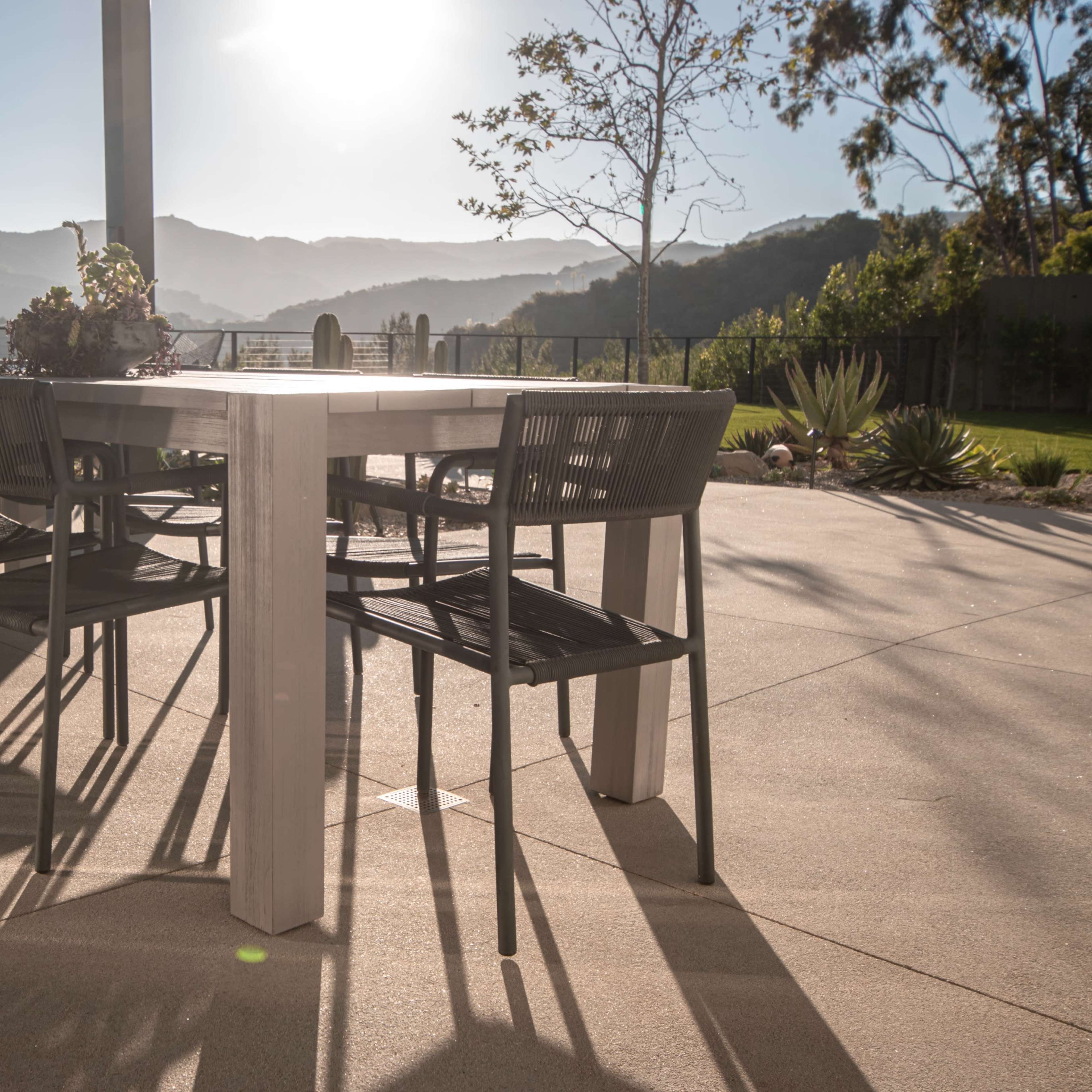 A dining table with chairs is set on a patio, surrounded by mountains and bathed in sunlight.