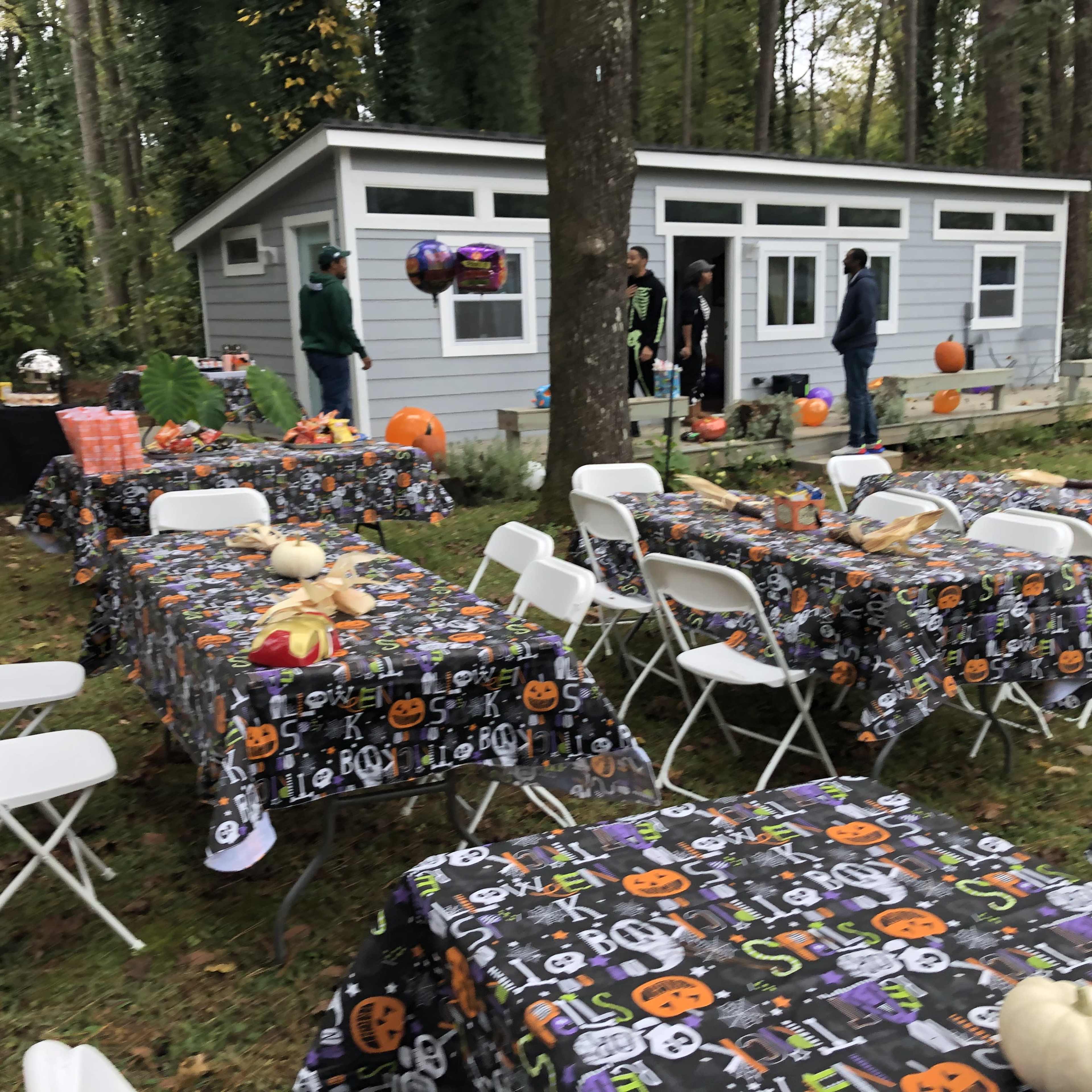 A backyard decorated for a Halloween party features tables with themed tablecloths, pumpkins, and guests socializing near a small gray house.