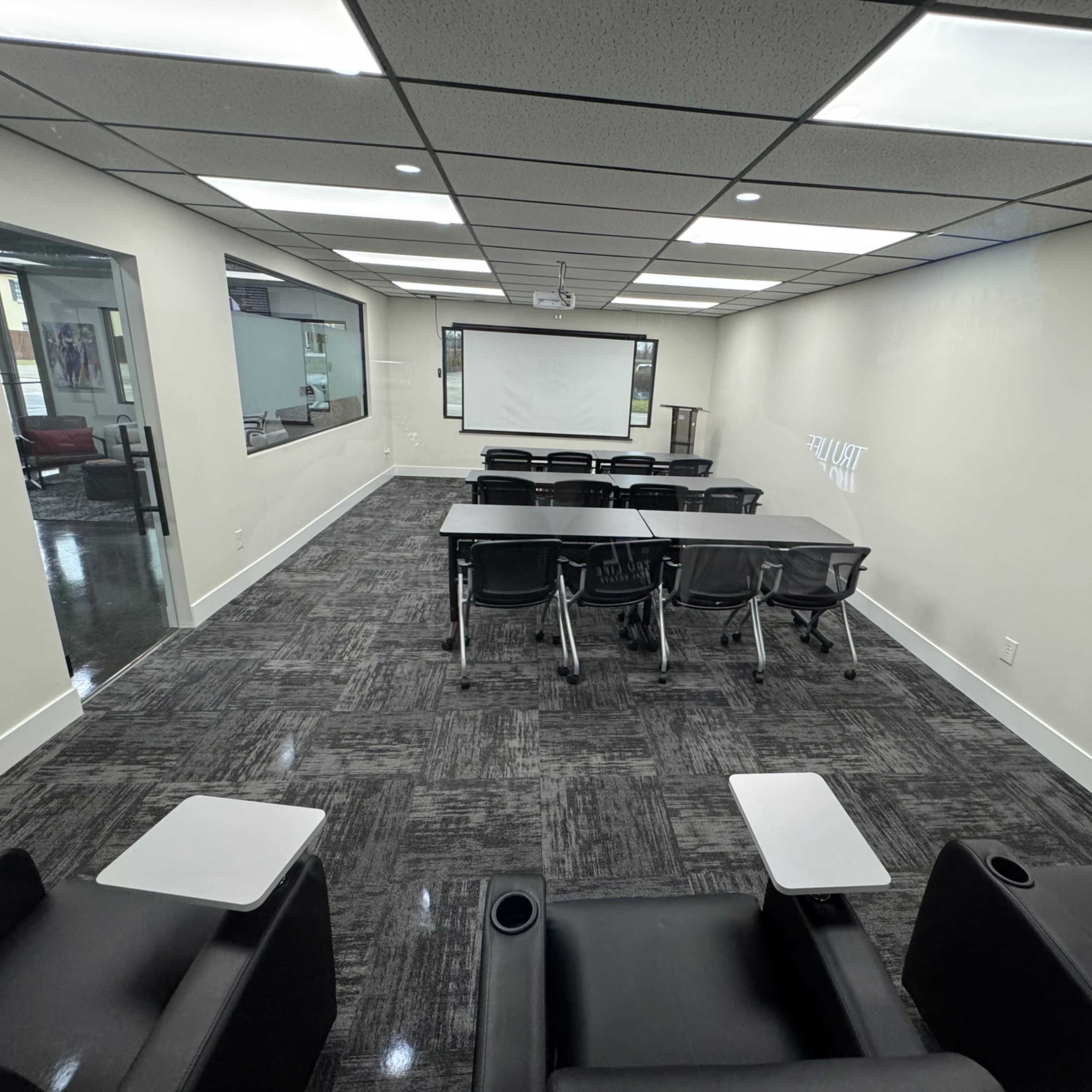 A modern meeting room features rows of tables and chairs, with a projector screen and a dark carpet.