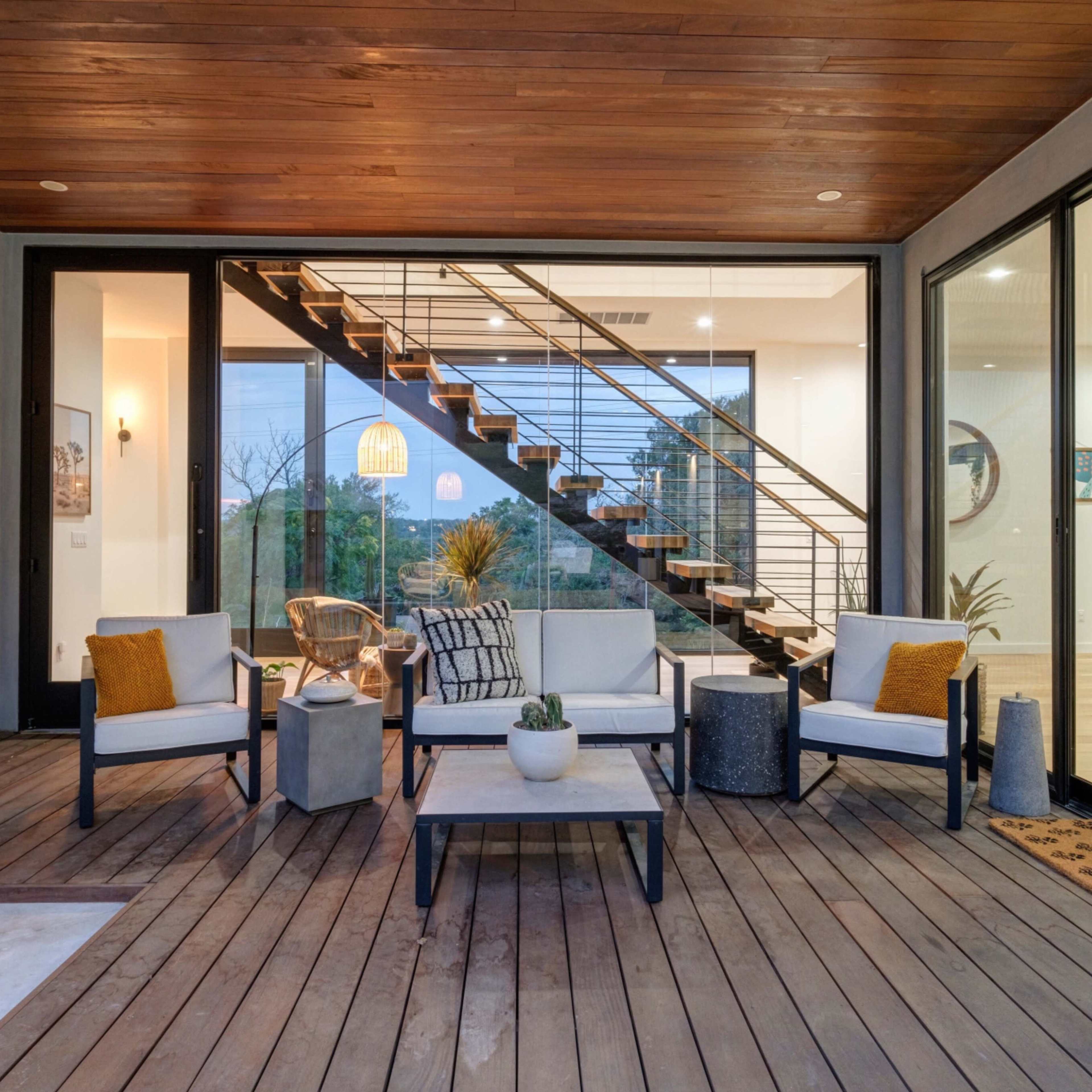 A modern outdoor seating area with a wooden deck, featuring four chairs, a coffee table, and a staircase leading to an interior space with large glass windows.