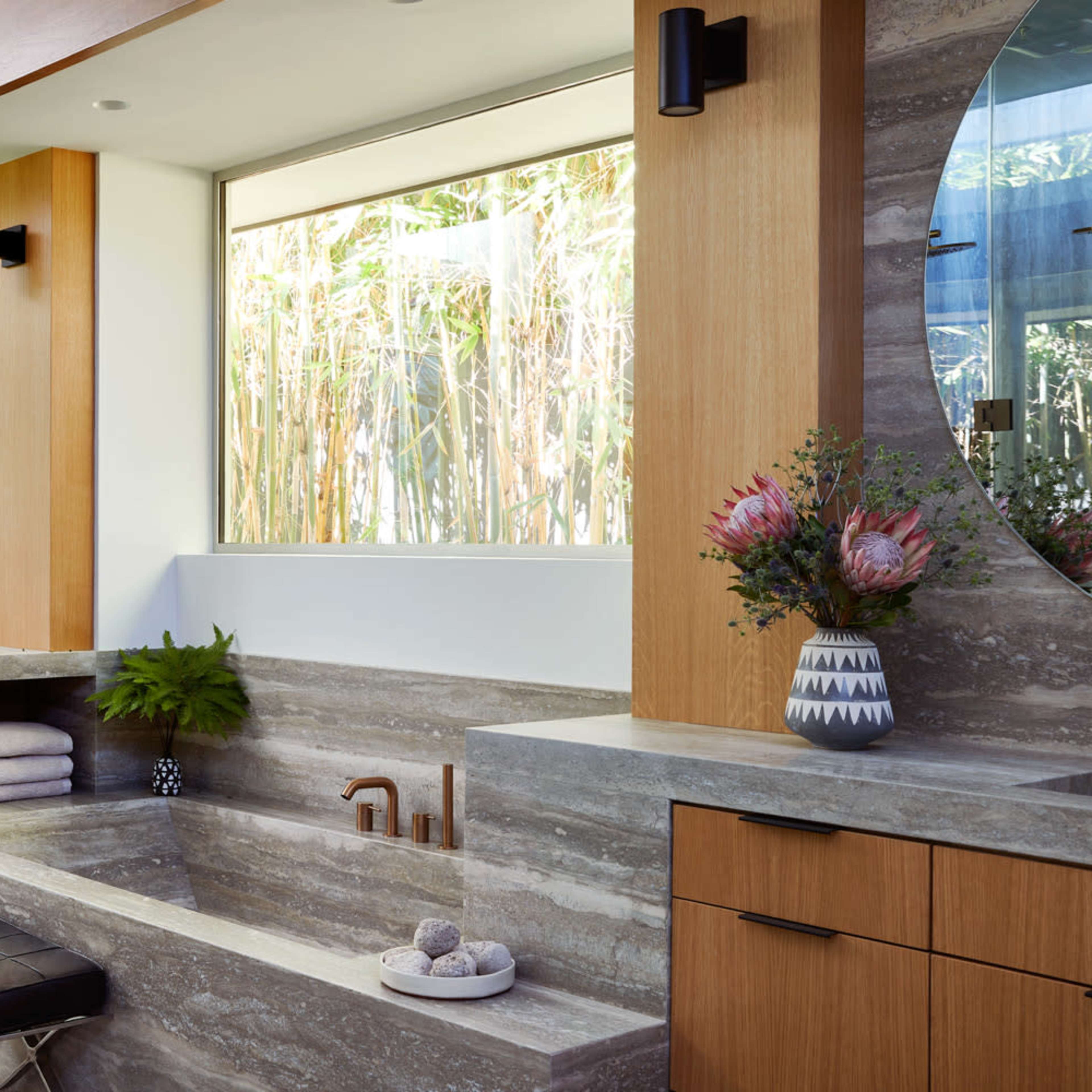 A modern bathroom with a stone bathtub, wooden cabinetry, large round mirrors, and large windows providing natural light.