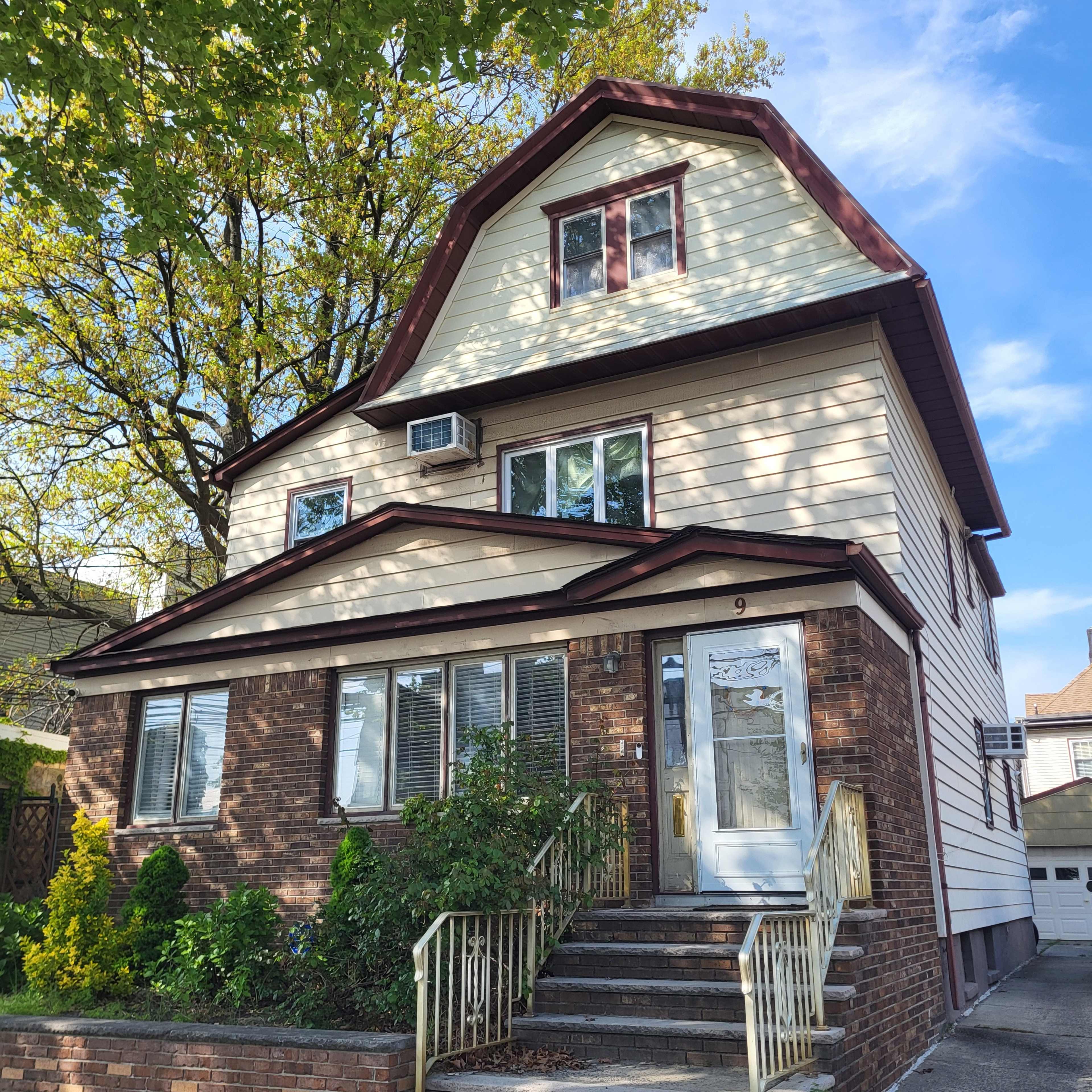 A three-story house with a gabled roof features a mix of brick and siding exterior, an air conditioning unit, and a front staircase with a railing.