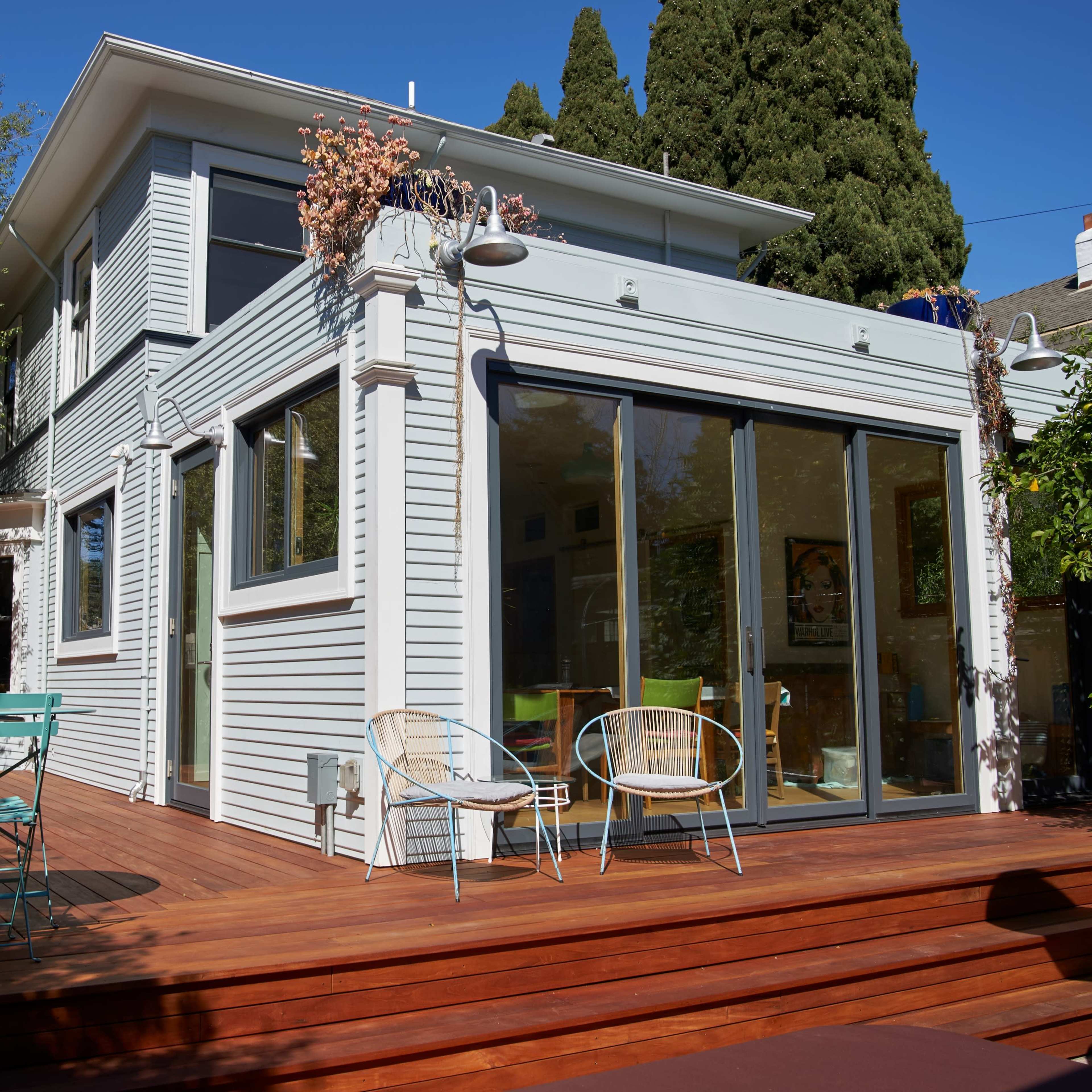 A modern house with large glass doors leading to a wooden deck, surrounded by trees and outdoor furniture.
