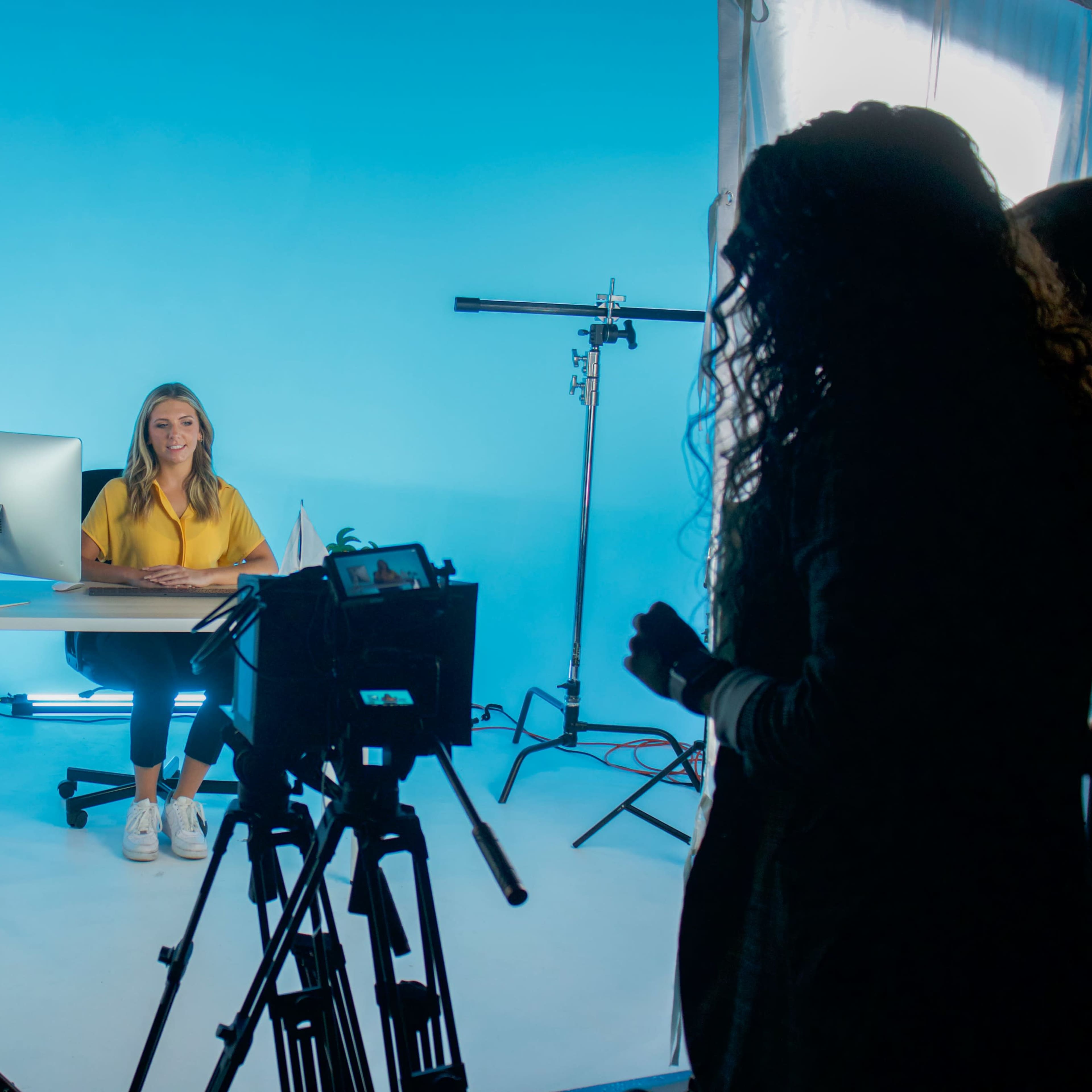 A woman sits at a desk in front of a computer, surrounded by studio lights and cameras with a blue backdrop.