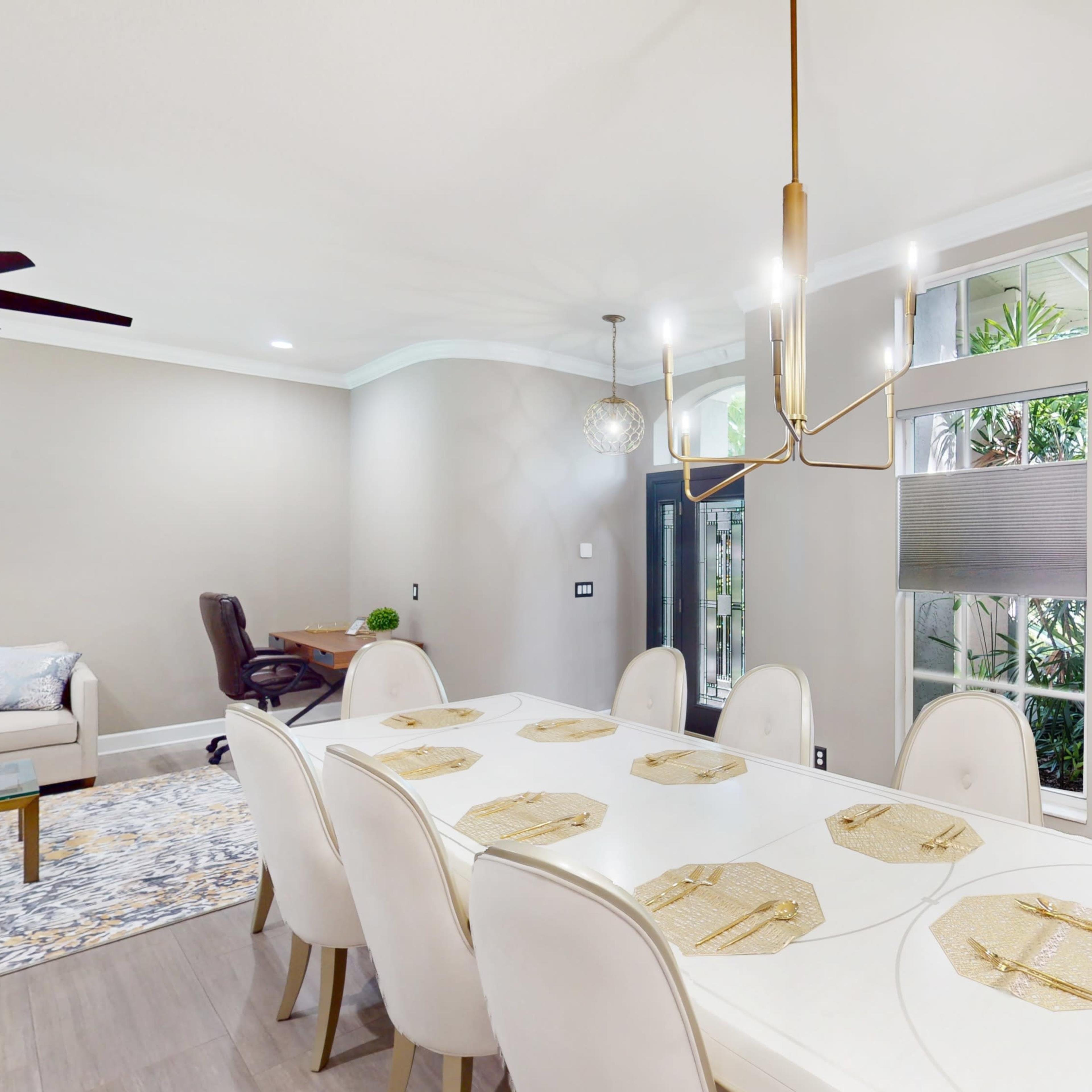 A modern dining area features a long white table with beige chairs, surrounded by large windows that provide natural light and views of greenery.