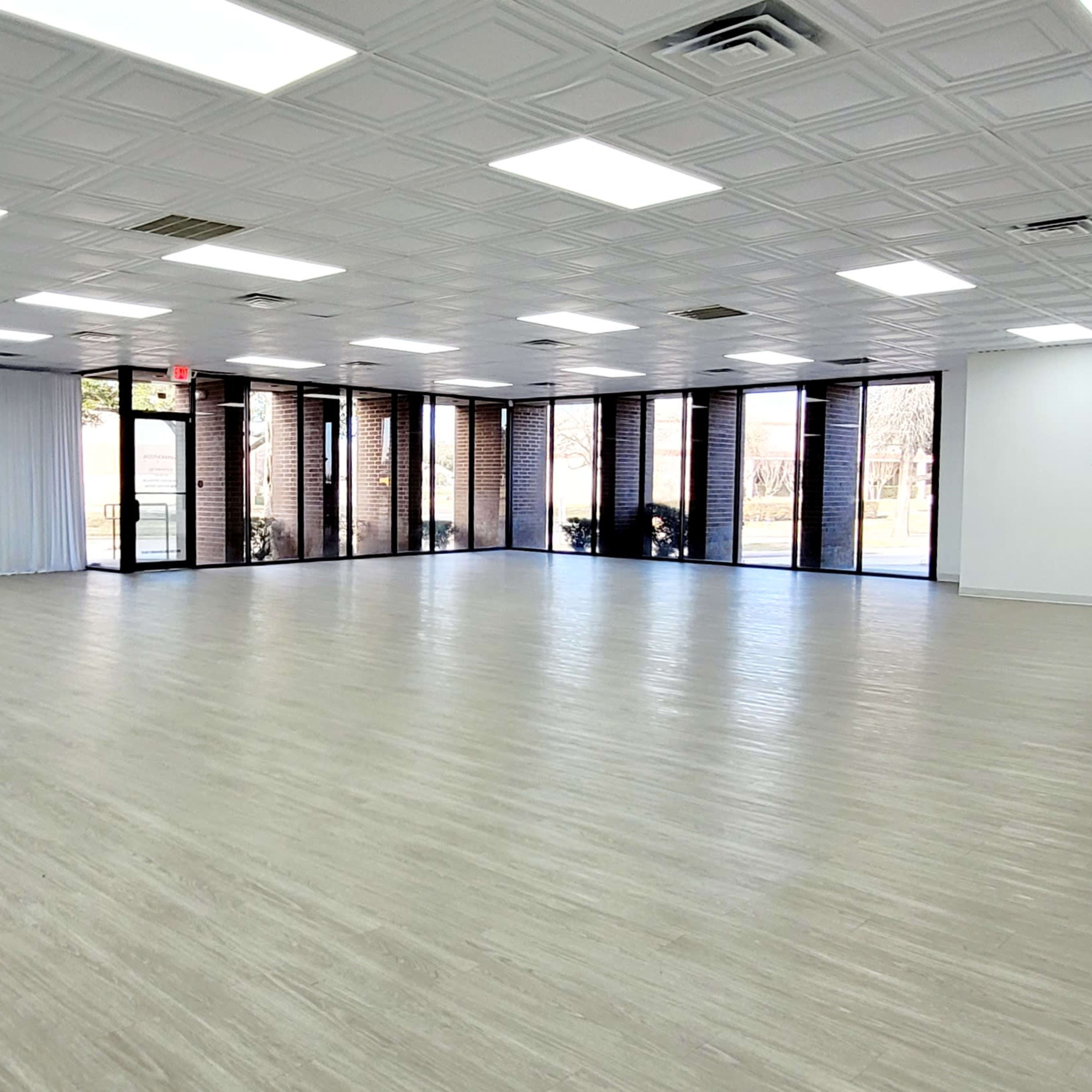 A spacious, empty interior room with a light wood floor, large windows, and white walls.