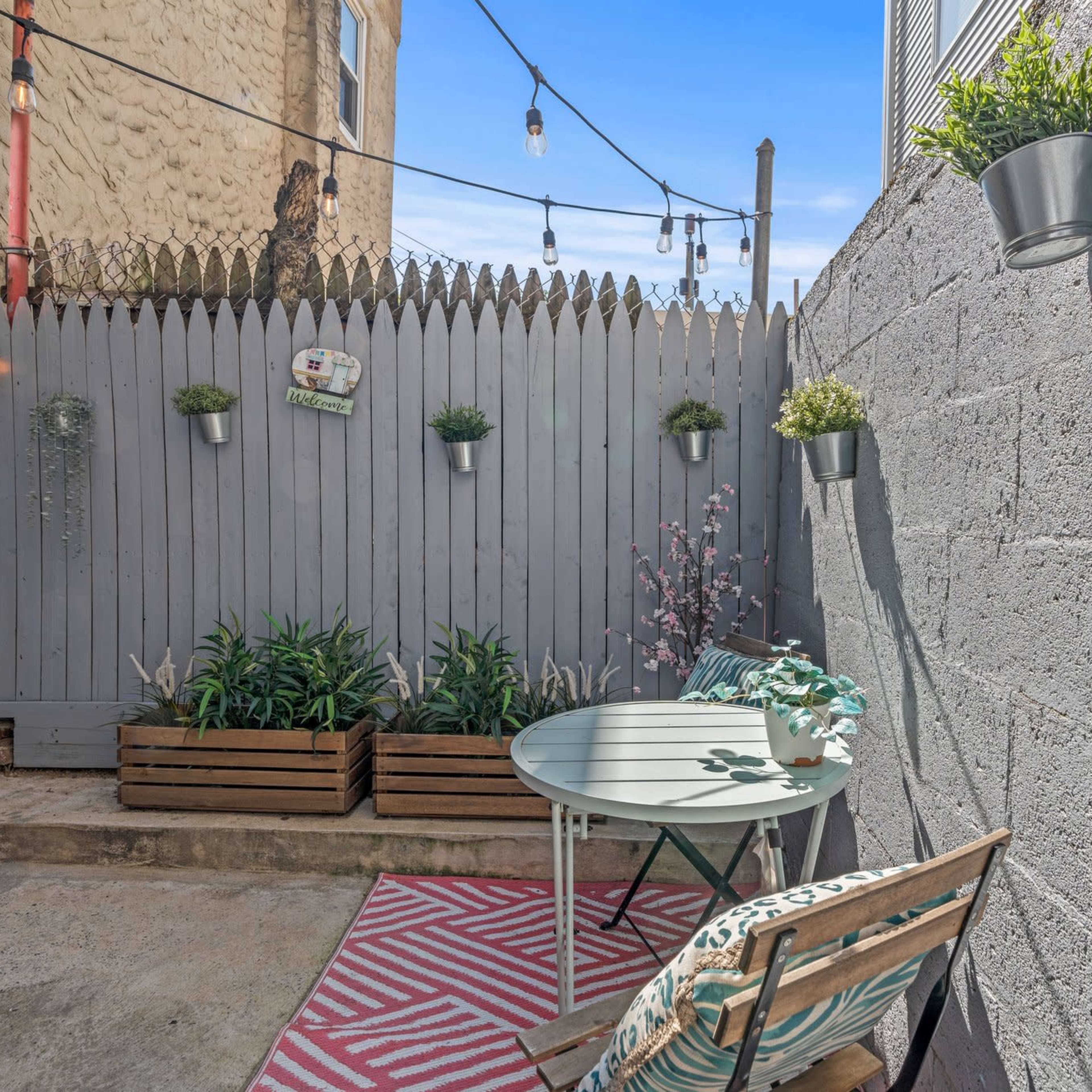 A small outdoor area with a table, a chair, potted plants, and string lights hanging above a gray wood fence.