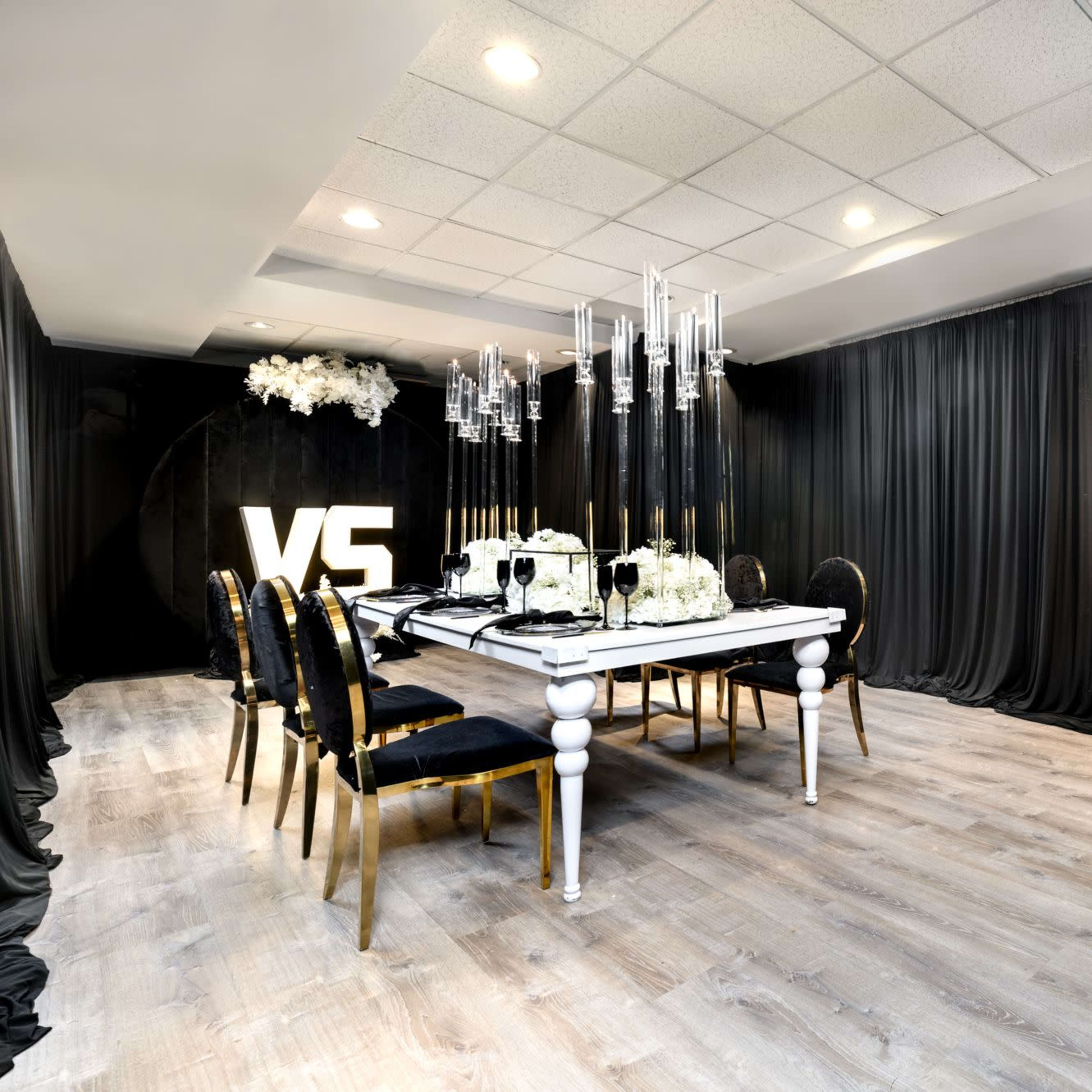 A dining room with a long table set for a meal, surrounded by black drapes and illuminated by a modern chandelier.