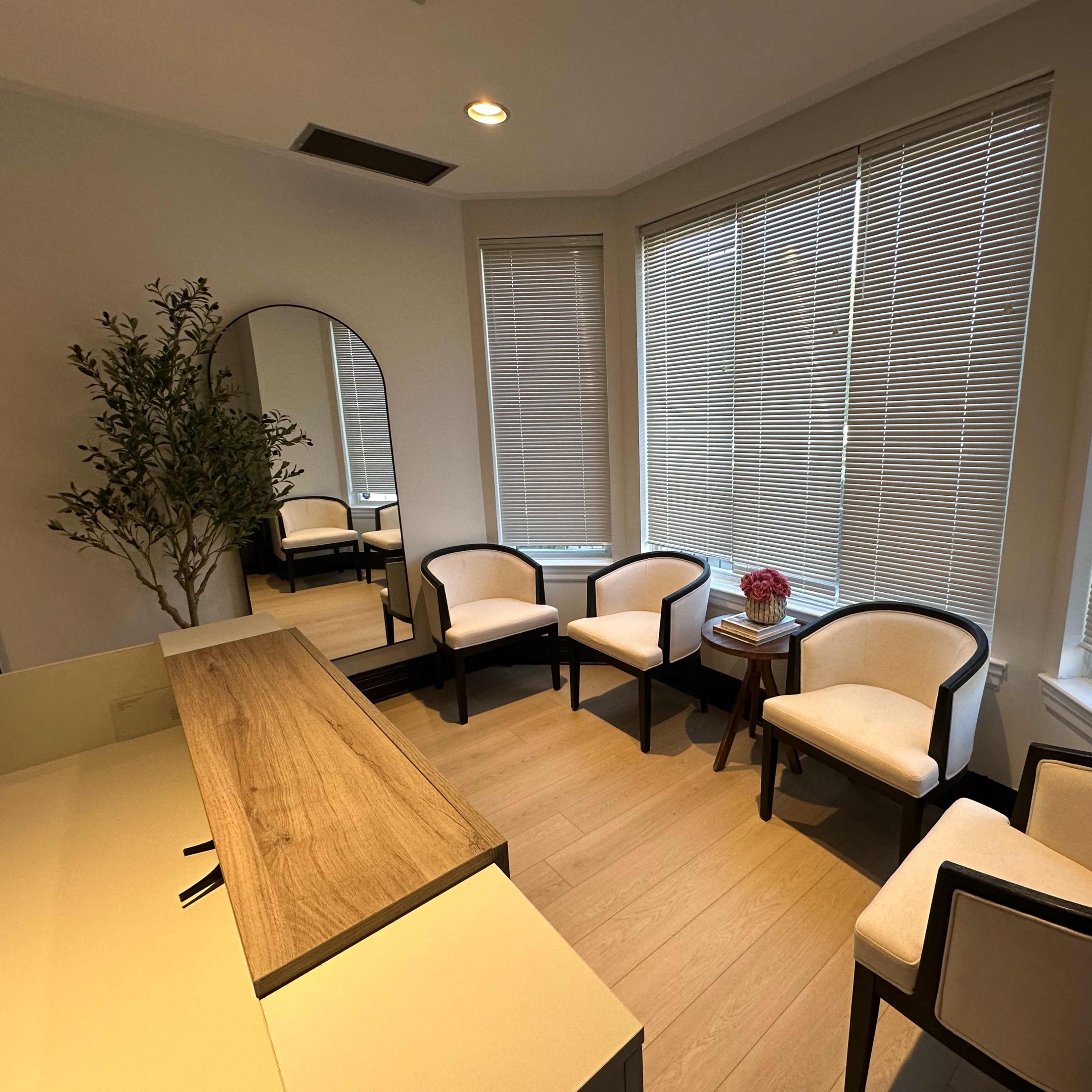 A small waiting room with four chairs, a mirror, a wooden table, and large windows covered by blinds.