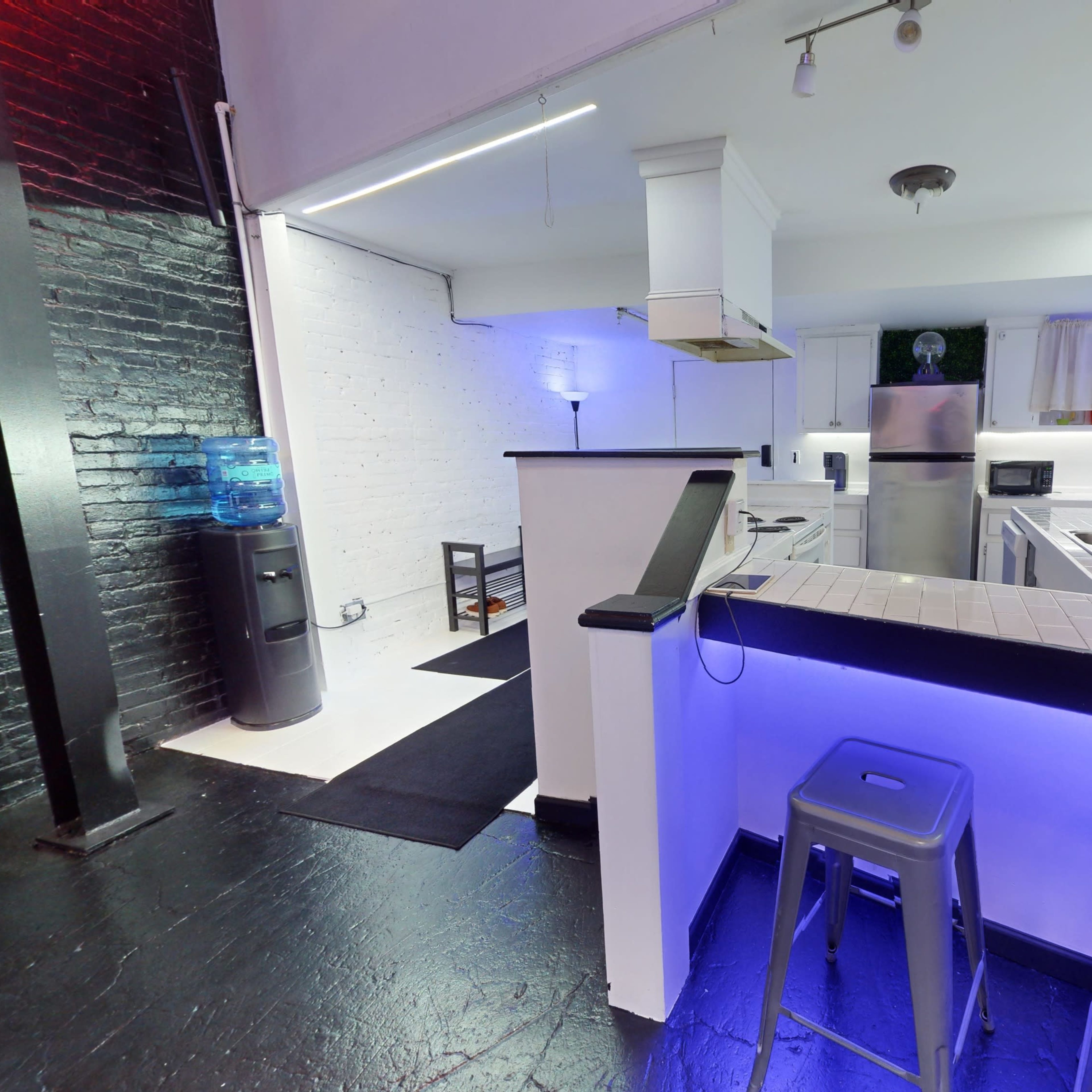A modern kitchen with stainless steel appliances, a white tile countertop, and blue accent lighting, adjacent to a black wall with a water cooler.