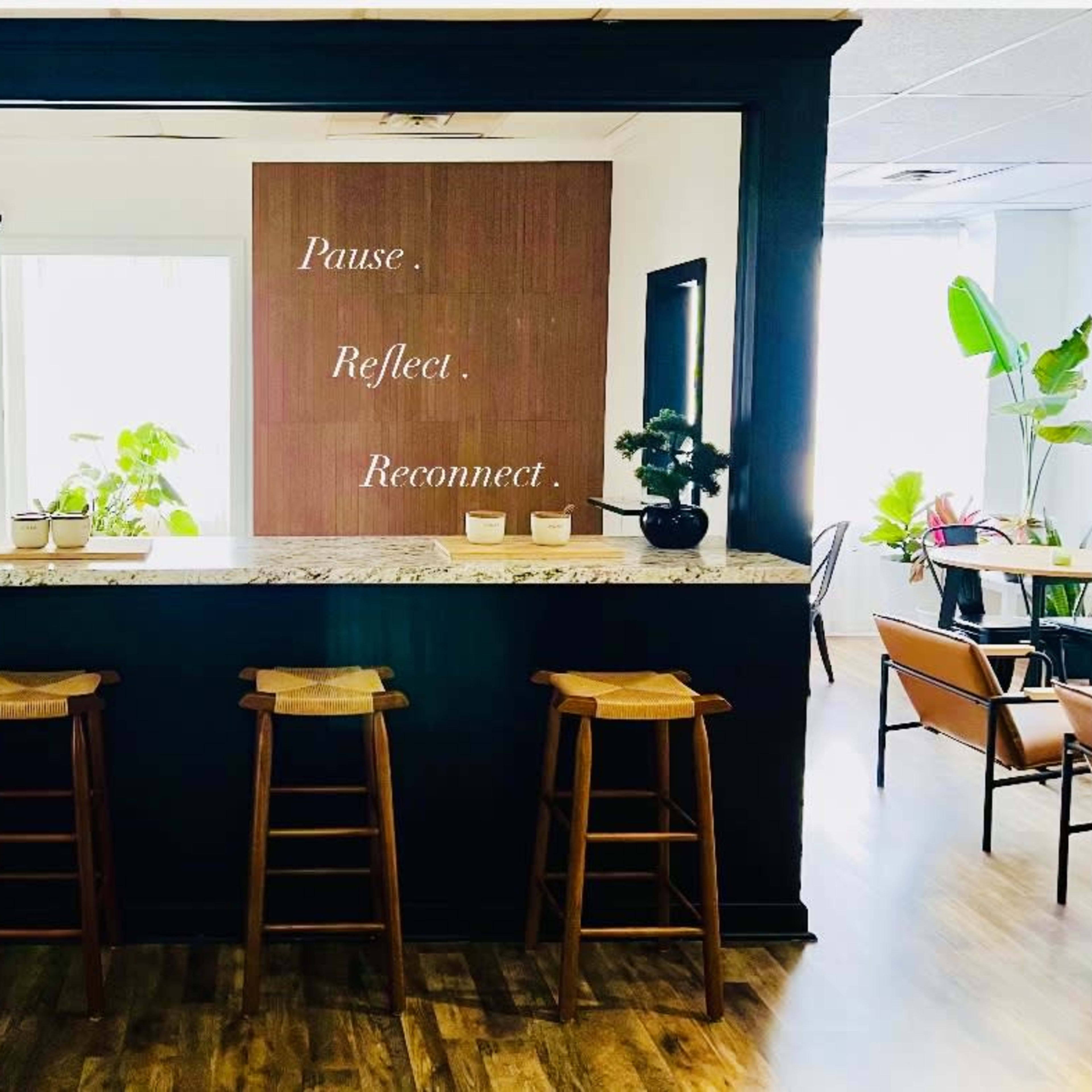 A minimalist reception area features a granite counter with three wooden stools and a wall displaying the words "Pause. Reflect. Reconnect."