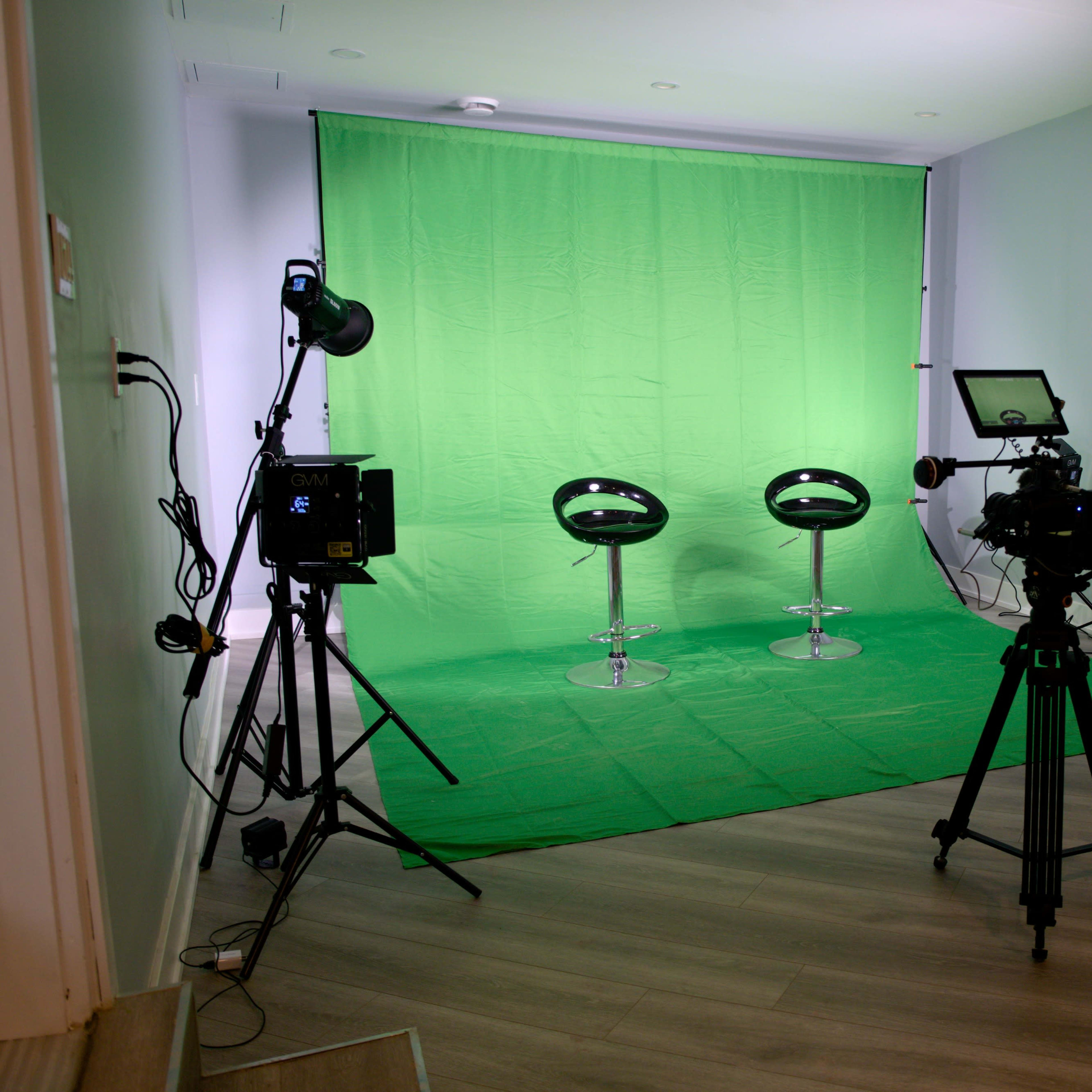 A photography studio with two black chairs on a green backdrop, illuminated by various lights and equipped with a camera on a tripod.