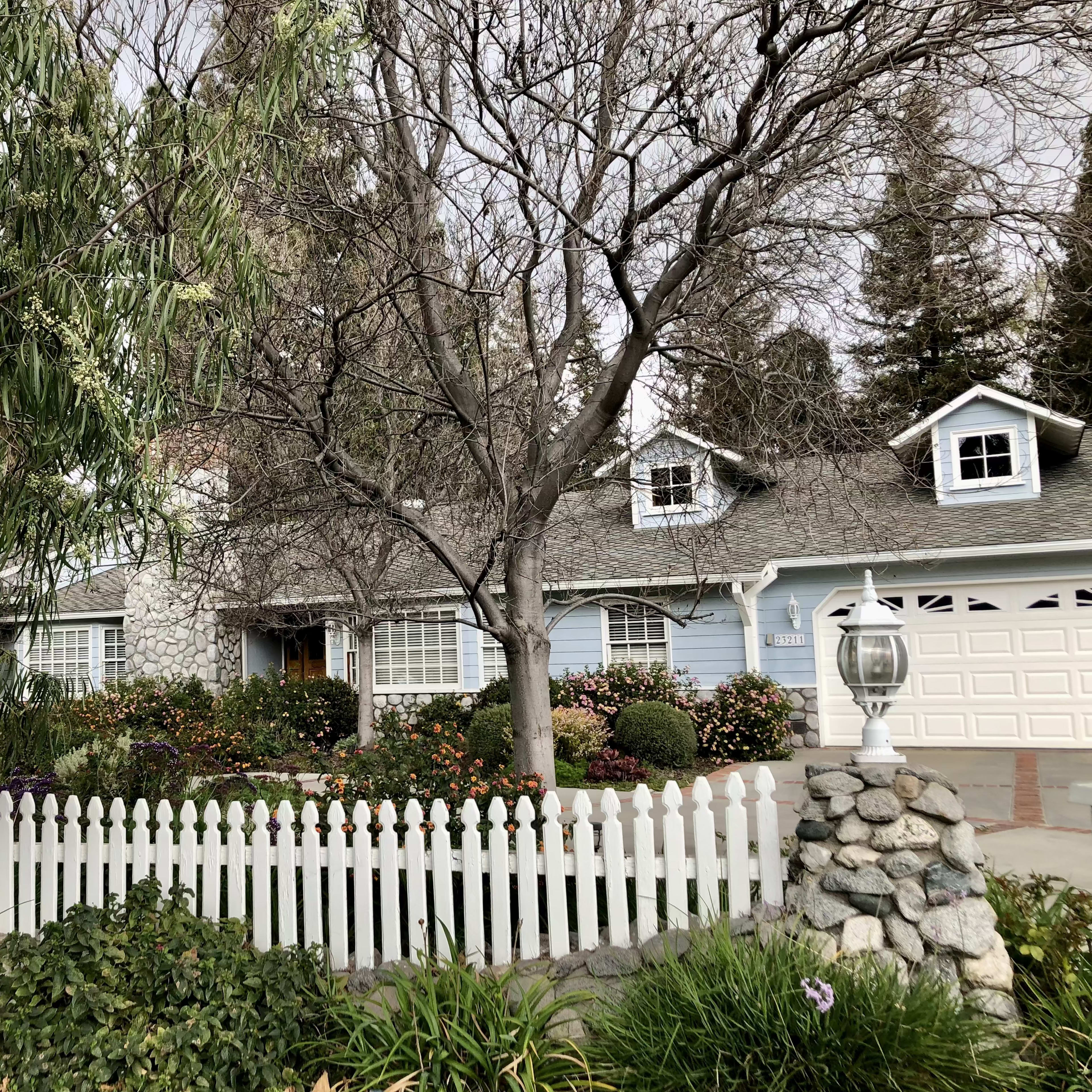 A house with a light blue exterior, surrounded by a white picket fence and various shrubs and trees, is visible in the scene.