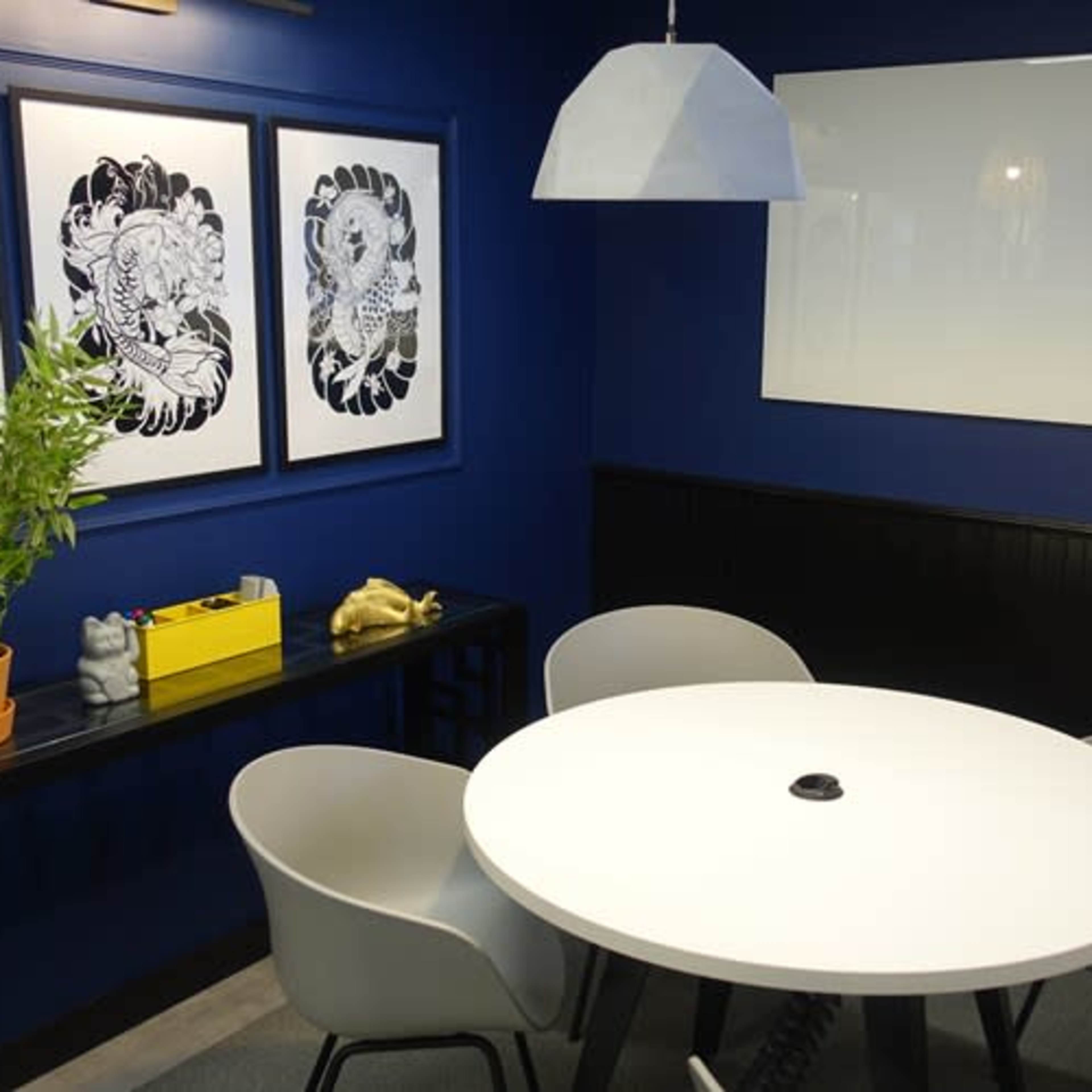 A modern meeting room featuring a round white table surrounded by four gray chairs, with dark blue walls adorned by two black-and-white framed artworks, and a hanging lamp above the table.