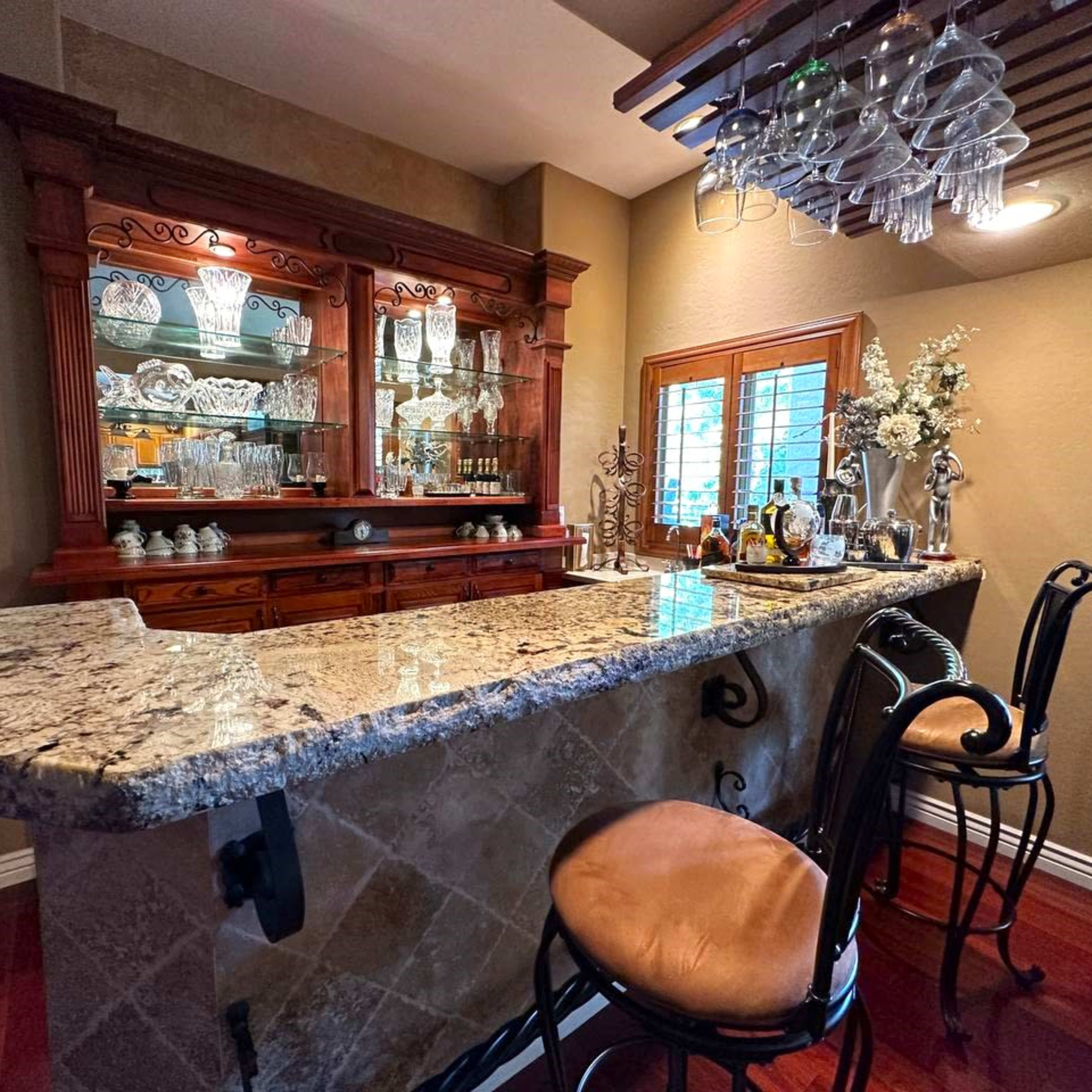A home bar area with a granite countertop, wooden cabinetry, and a display of glassware under a hanging light fixture.