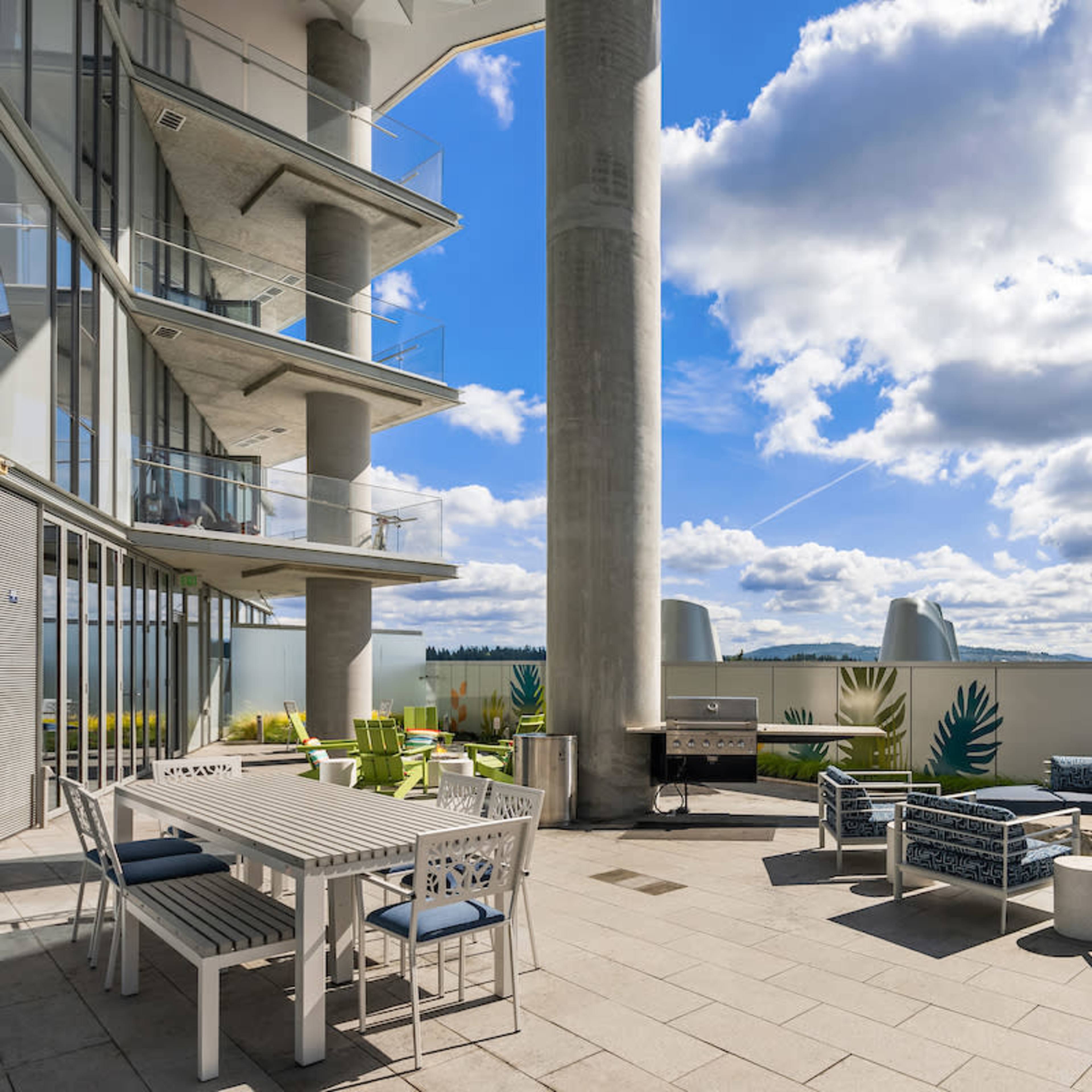 A modern balcony terrace with seating arrangements, a dining table, and a grill, framed by tall buildings and a blue sky filled with clouds.