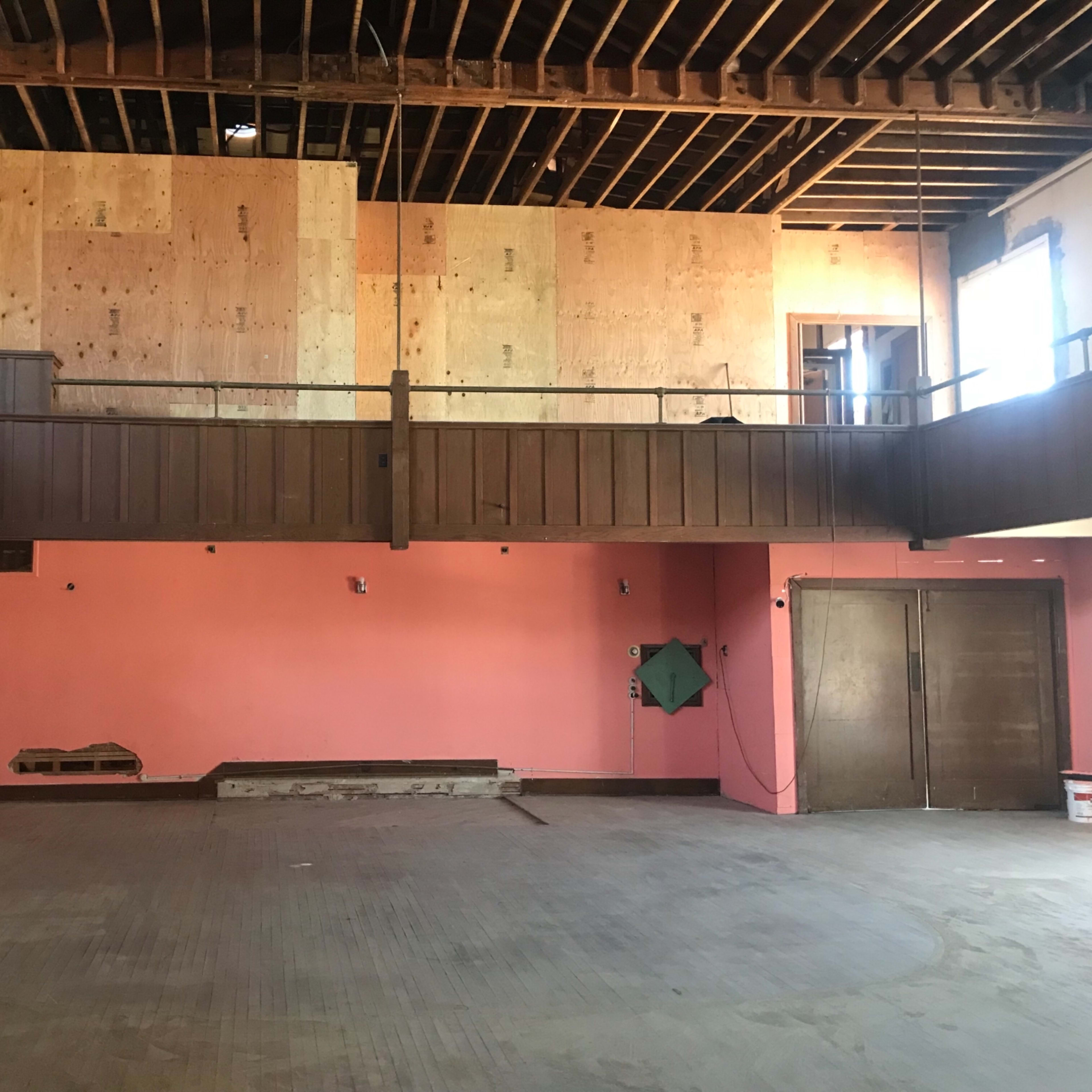 An unfinished interior space with exposed beams, plywood walls, and a pink-painted wall, featuring two doors and a balcony area.