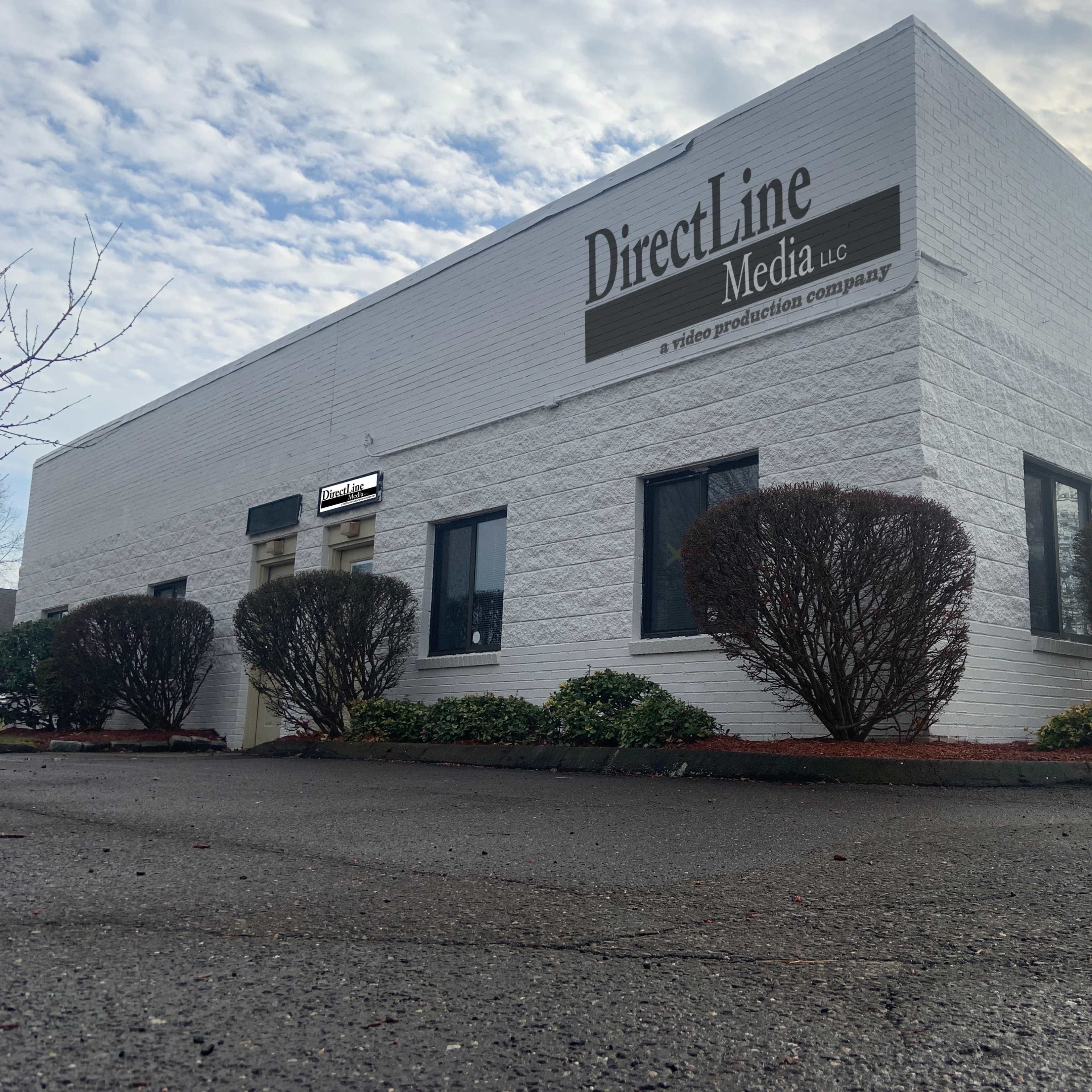 A one-story white building with "DirectLine Media" signage and landscaping, situated on a paved area under a cloudy sky.