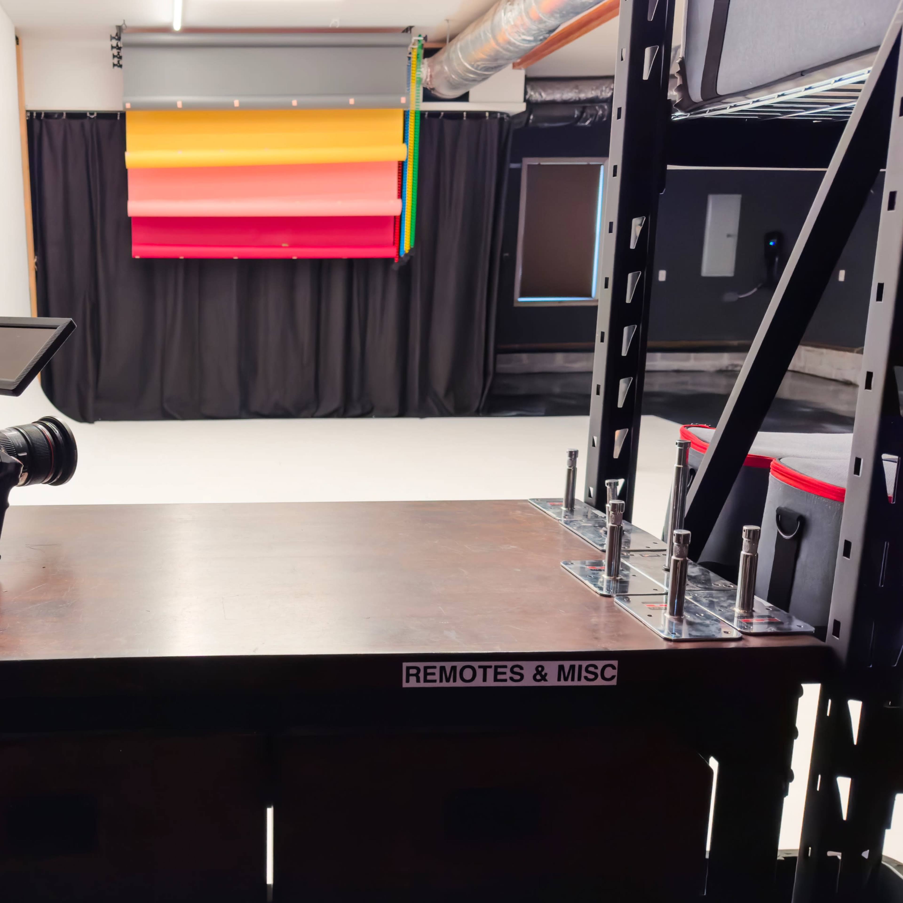 A camera is positioned on a wooden table labeled "MAIN LENSES," facing a backdrop with colorful horizontal panels in a studio setting.