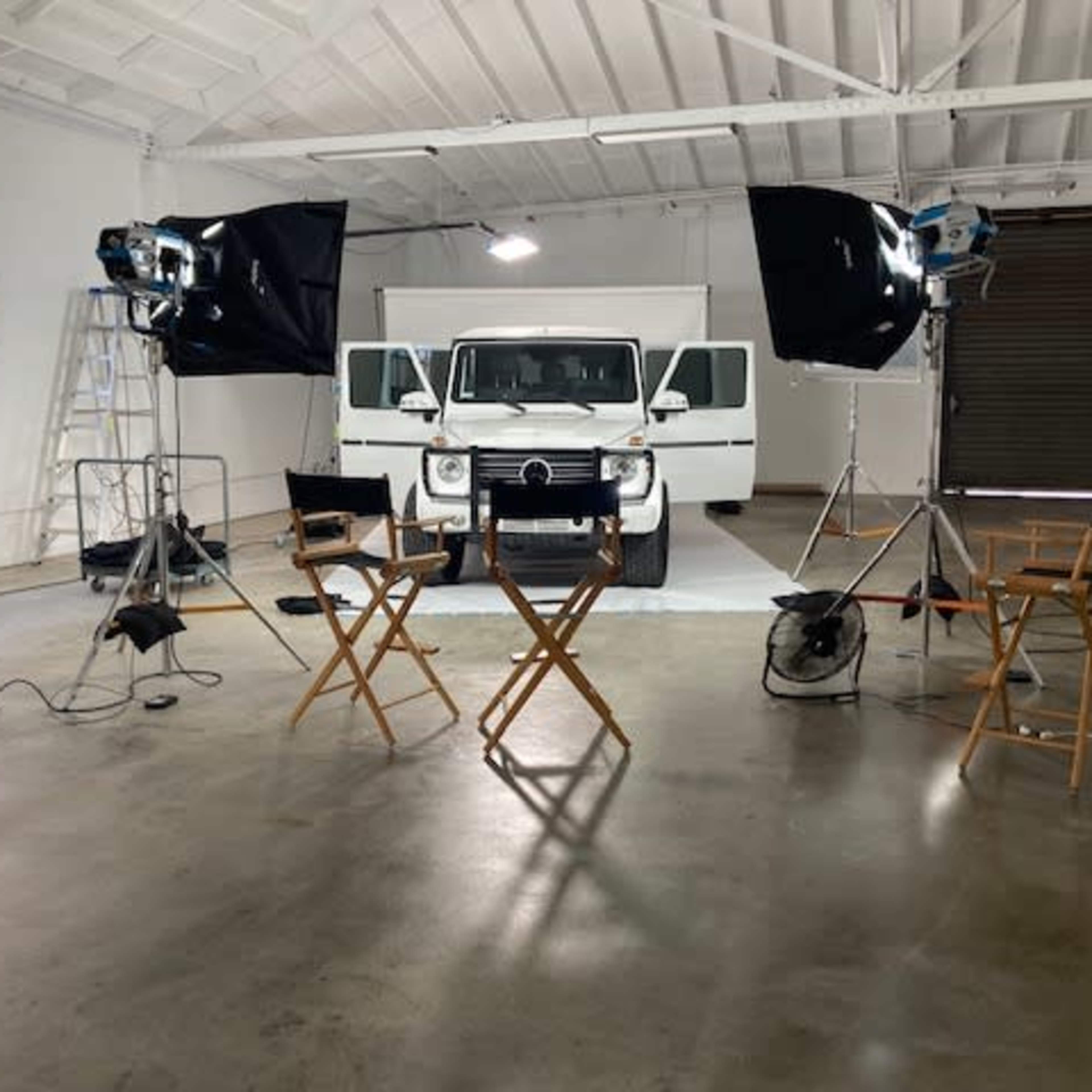 A studio setup with lights, cameras, and two director's chairs facing a white vehicle positioned in front of a backdrop.