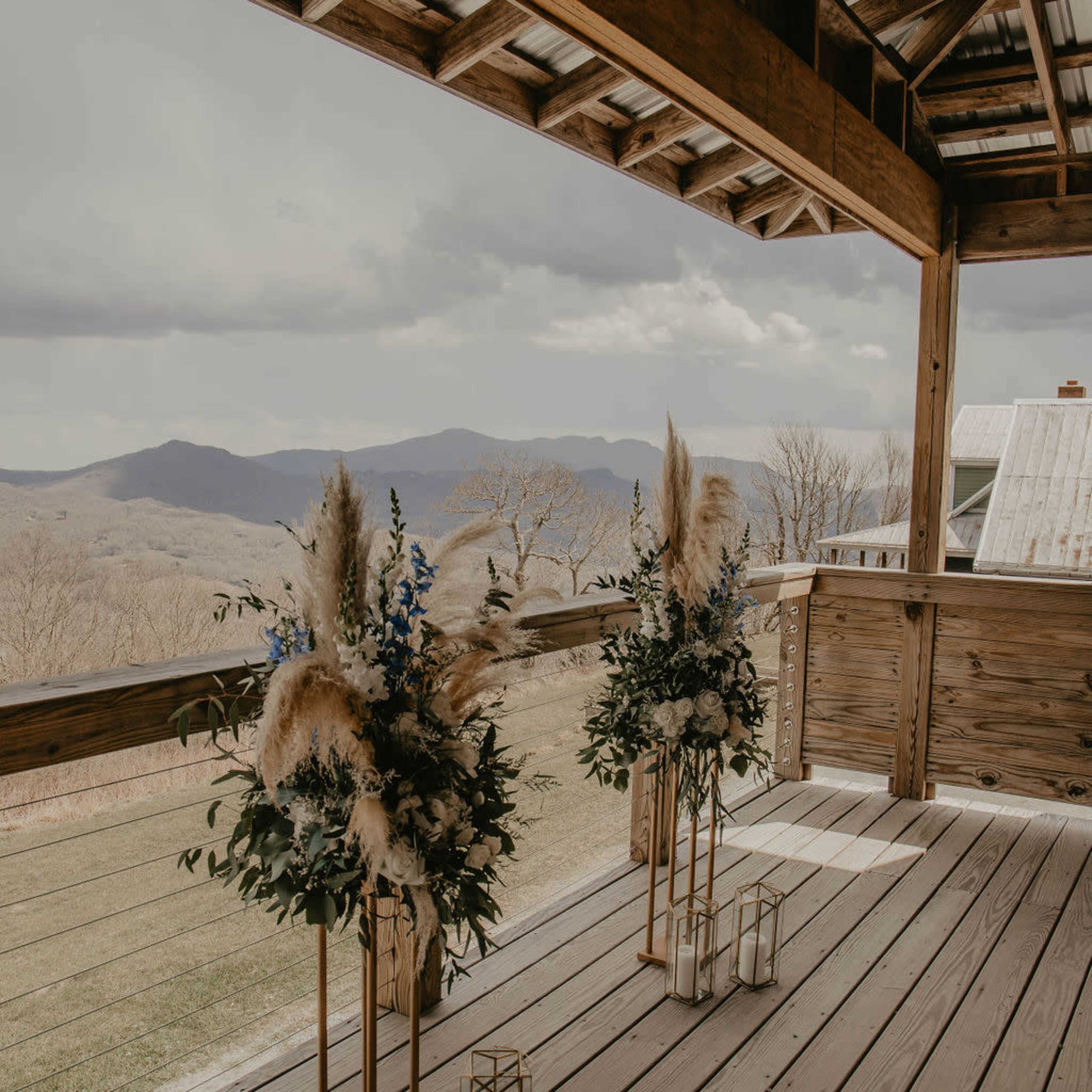 Two floral arrangements with blue flowers and pampas grass stand on a wooden deck overlooking a mountainous landscape under a cloudy sky.