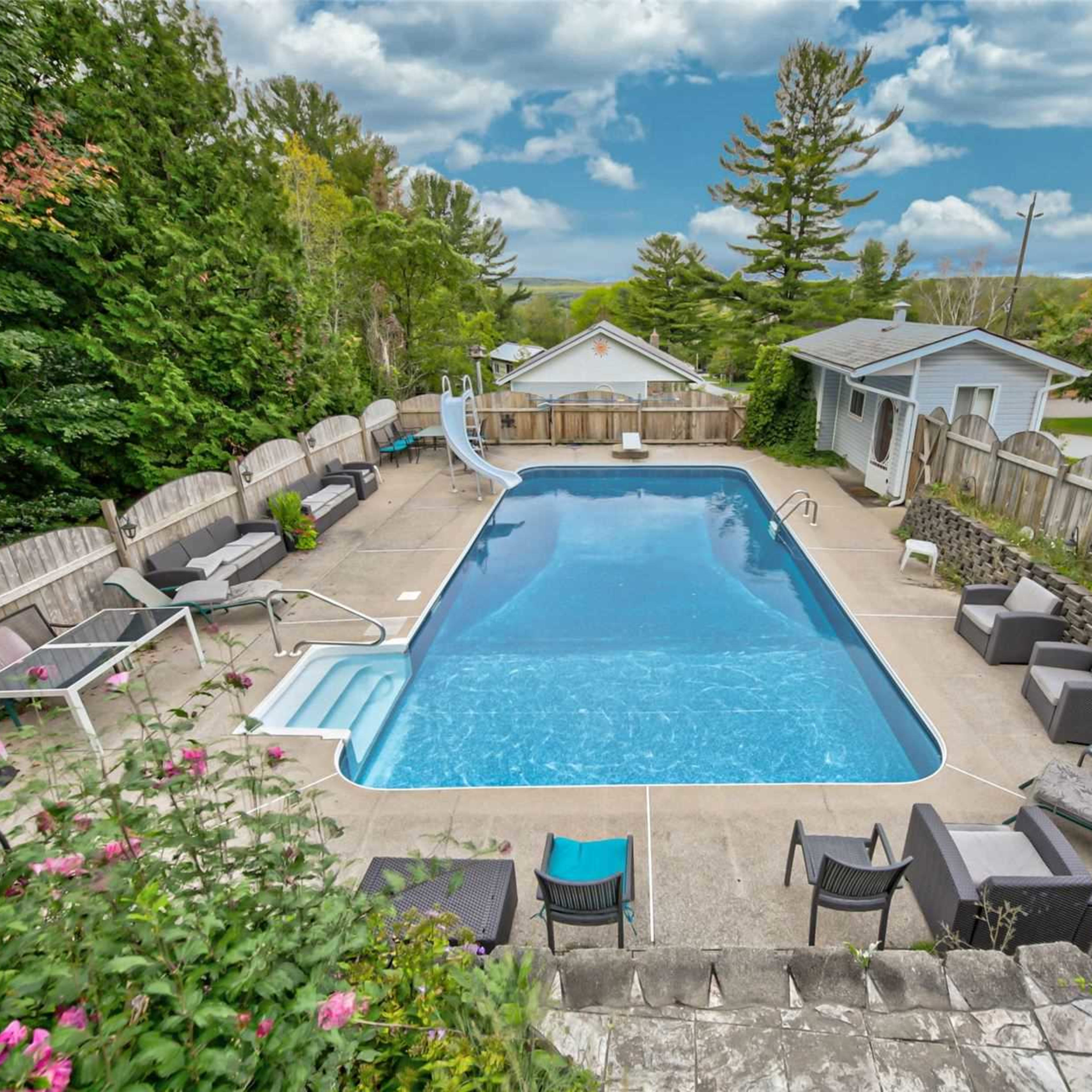 A rectangular swimming pool surrounded by lounge chairs and a slide, with a backdrop of trees and a house.