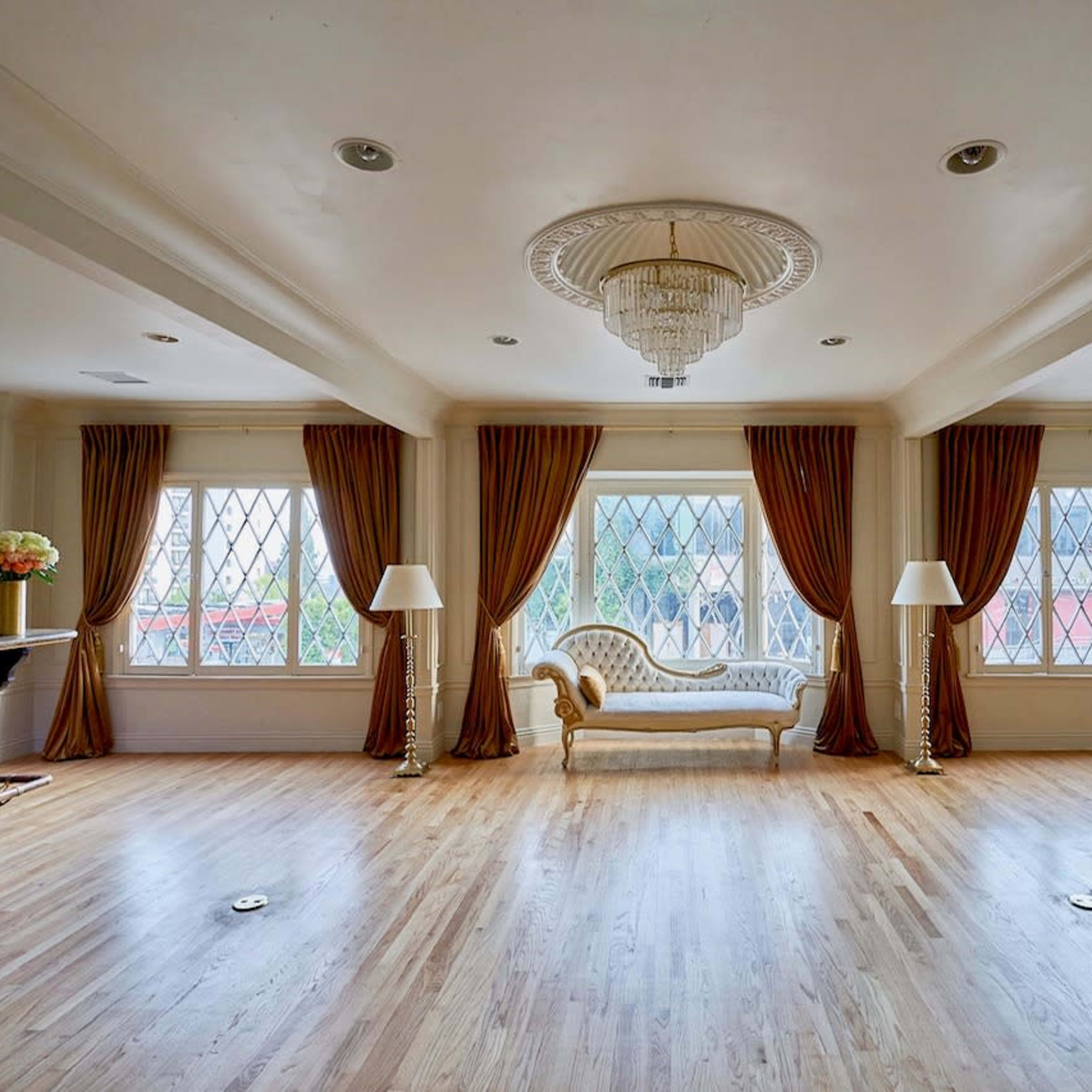 The room features a central chaise lounge flanked by two large windows draped in curtains, with a chandelier overhead and lamps at each end.