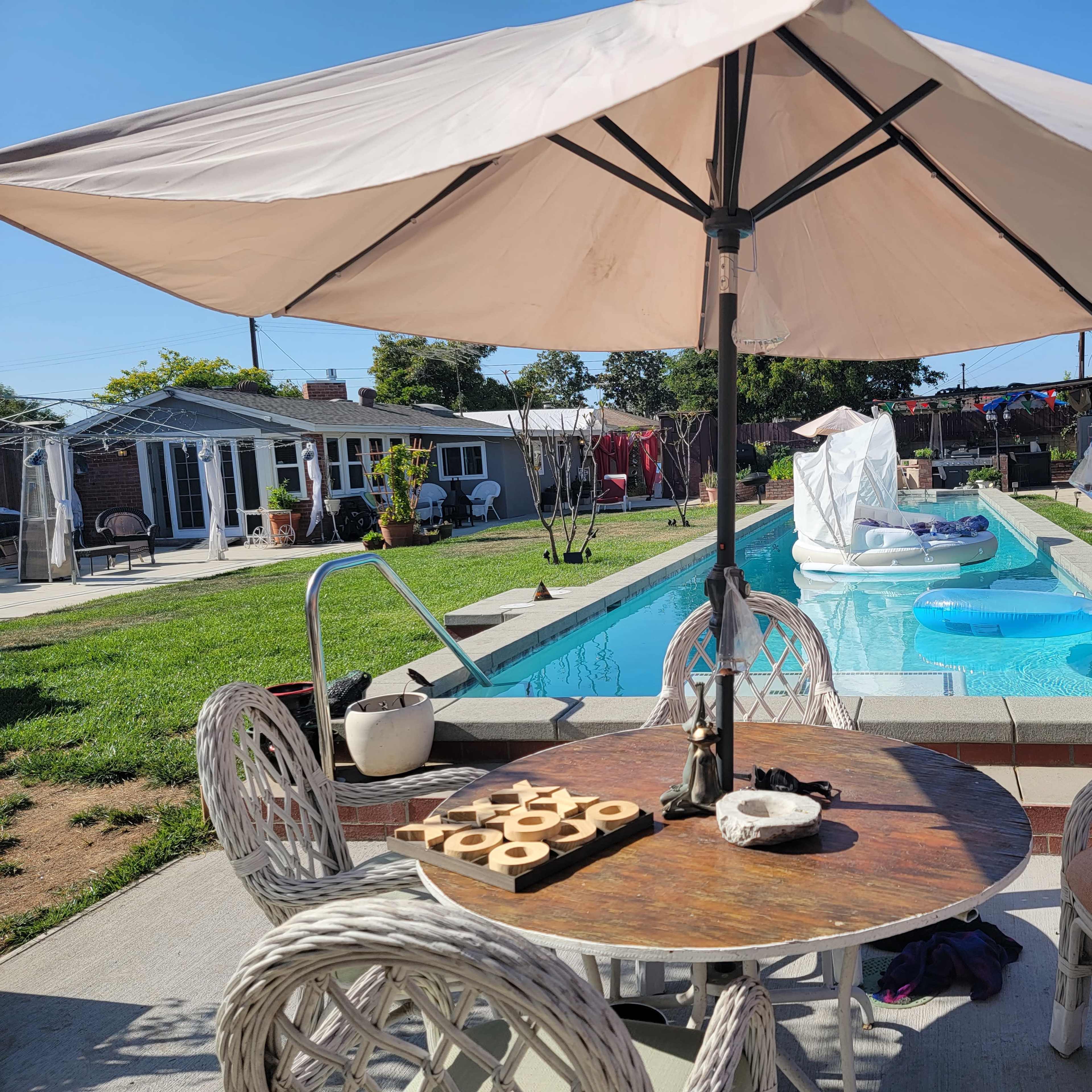 A backyard pool area with a table and chairs under an umbrella, a clean pool with inflatable toys, and various outdoor structures in the background.