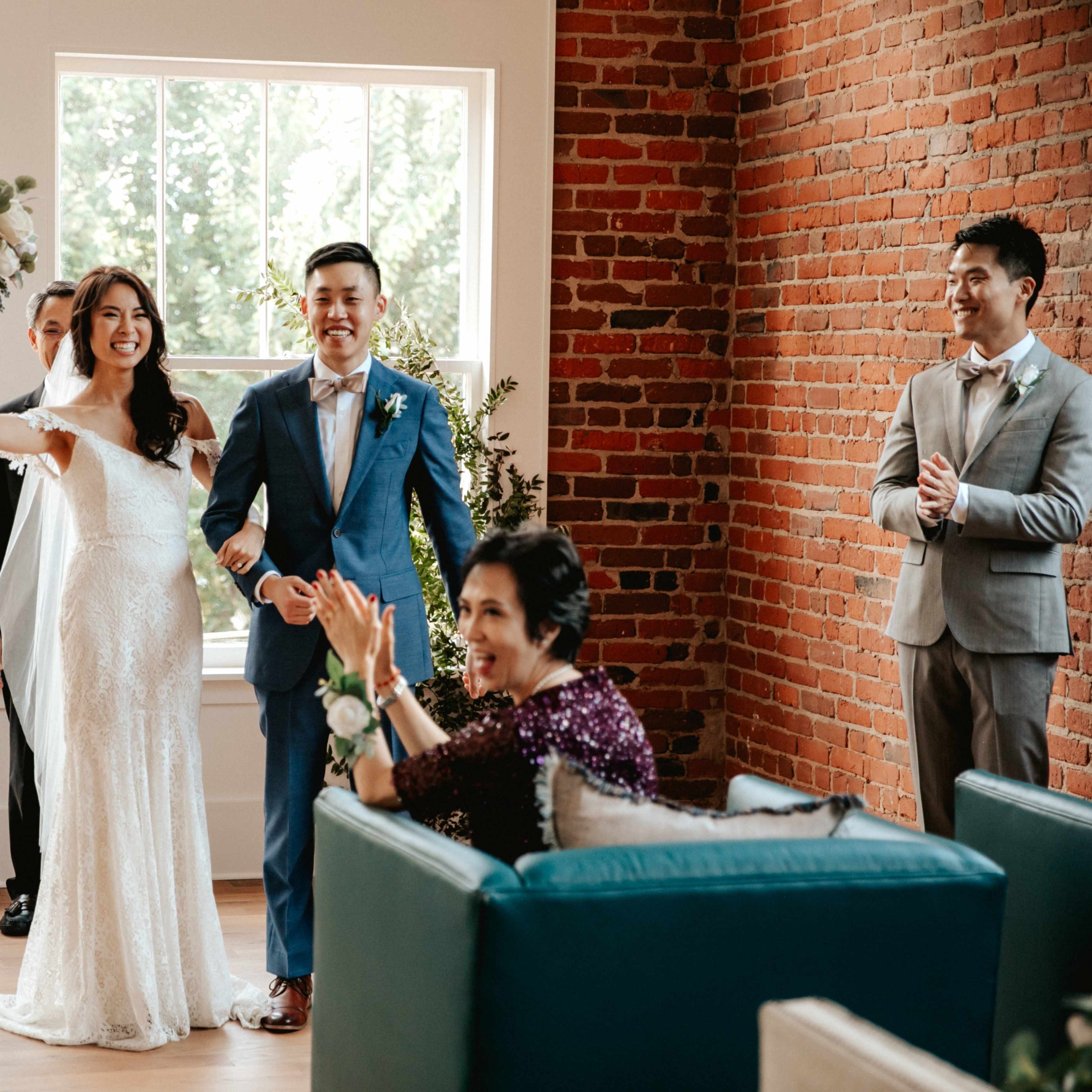 A bride in a wedding gown and a groom in a suit stand together, smiling and celebrating with family and friends in a room with brick walls and large windows.