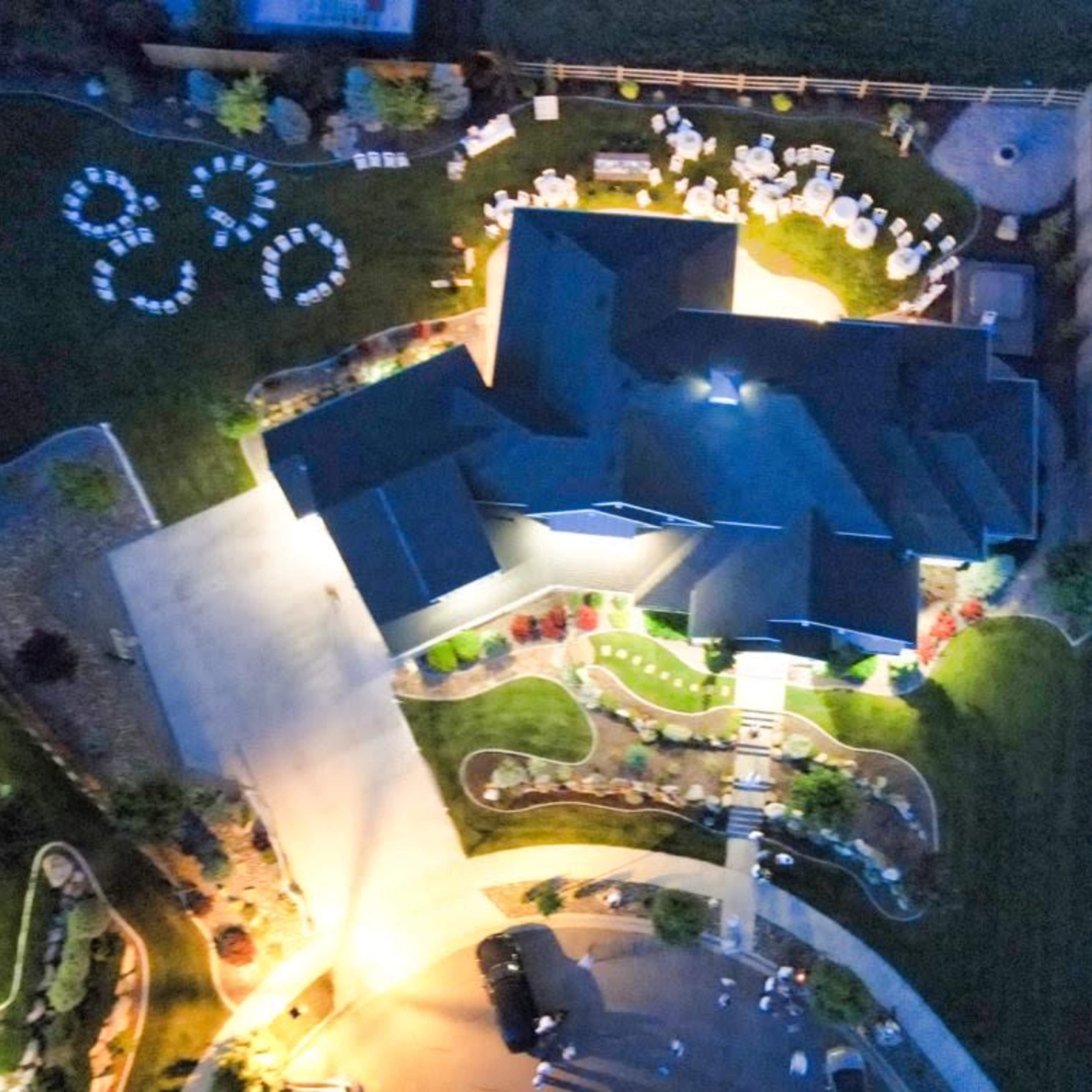 An aerial view shows a large house surrounded by landscaped gardens and seating arranged for an event in the yard.