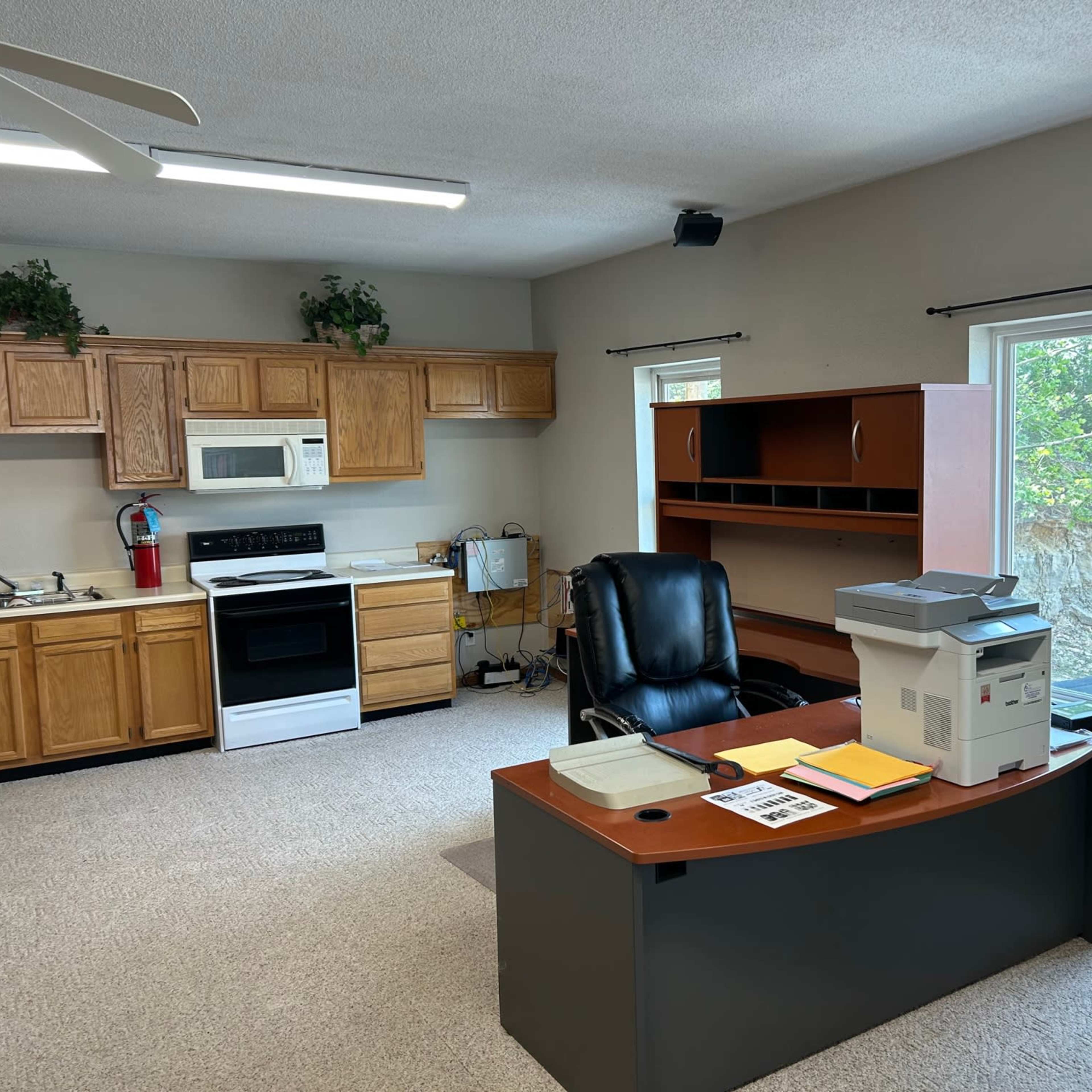 A small office space with a wooden desk, black chair, a kitchen area with appliances, and a window overlooking greenery outside.