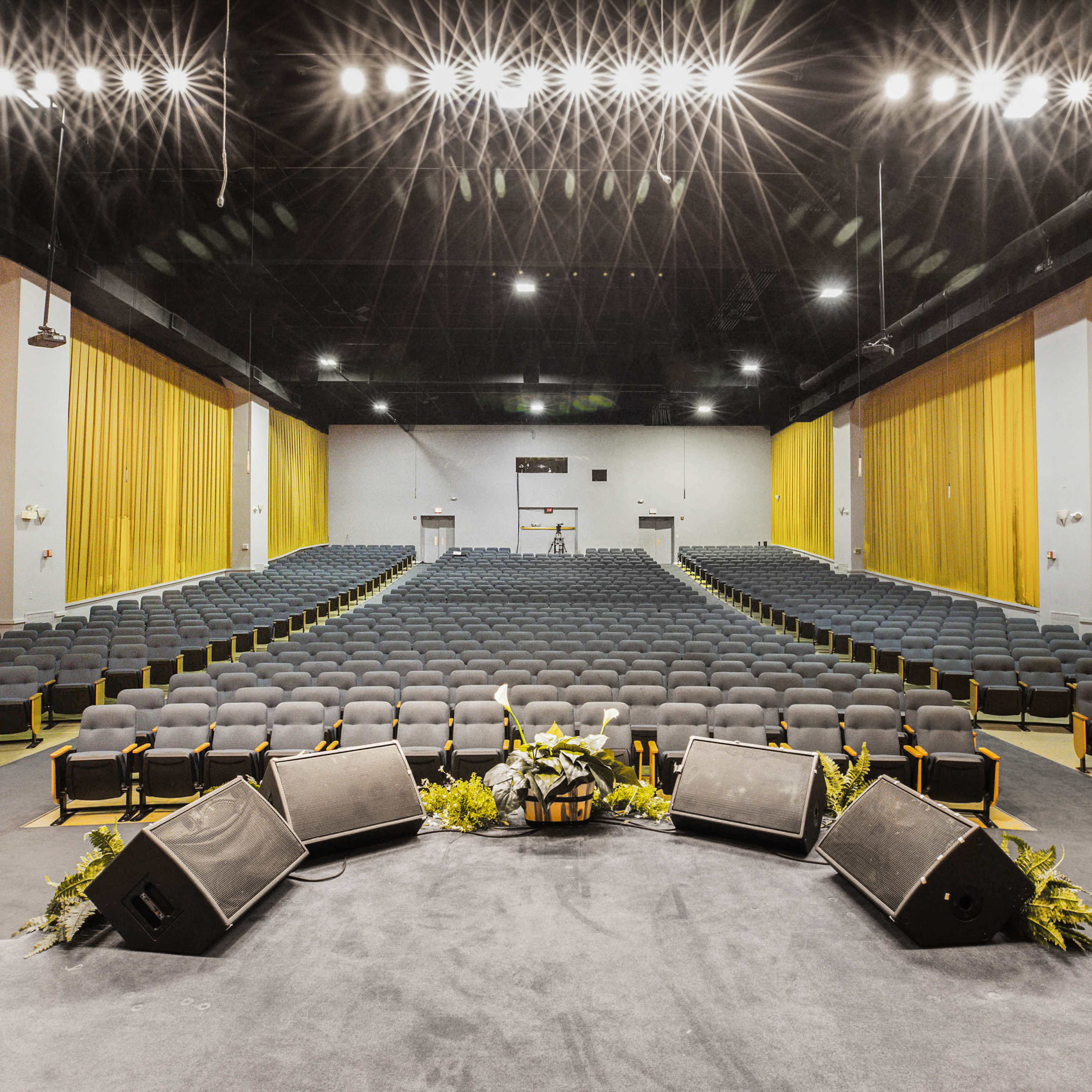 An empty auditorium with rows of black chairs and a stage at the forefront, surrounded by yellow curtains and brightly lit from above.
