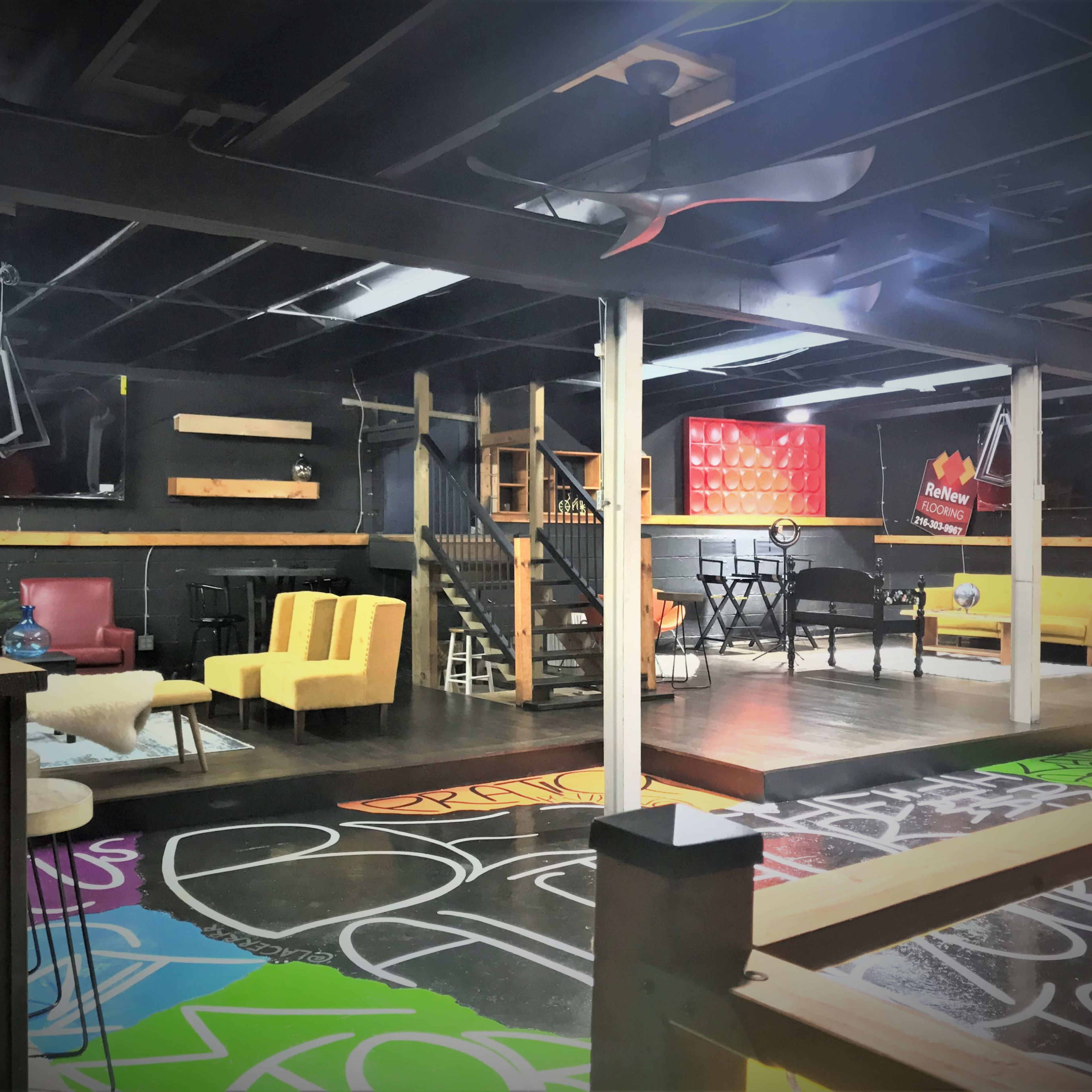 A spacious, colorful workshop or creative space featuring various seating areas, a staircase, and walls adorned with vibrant graffiti.