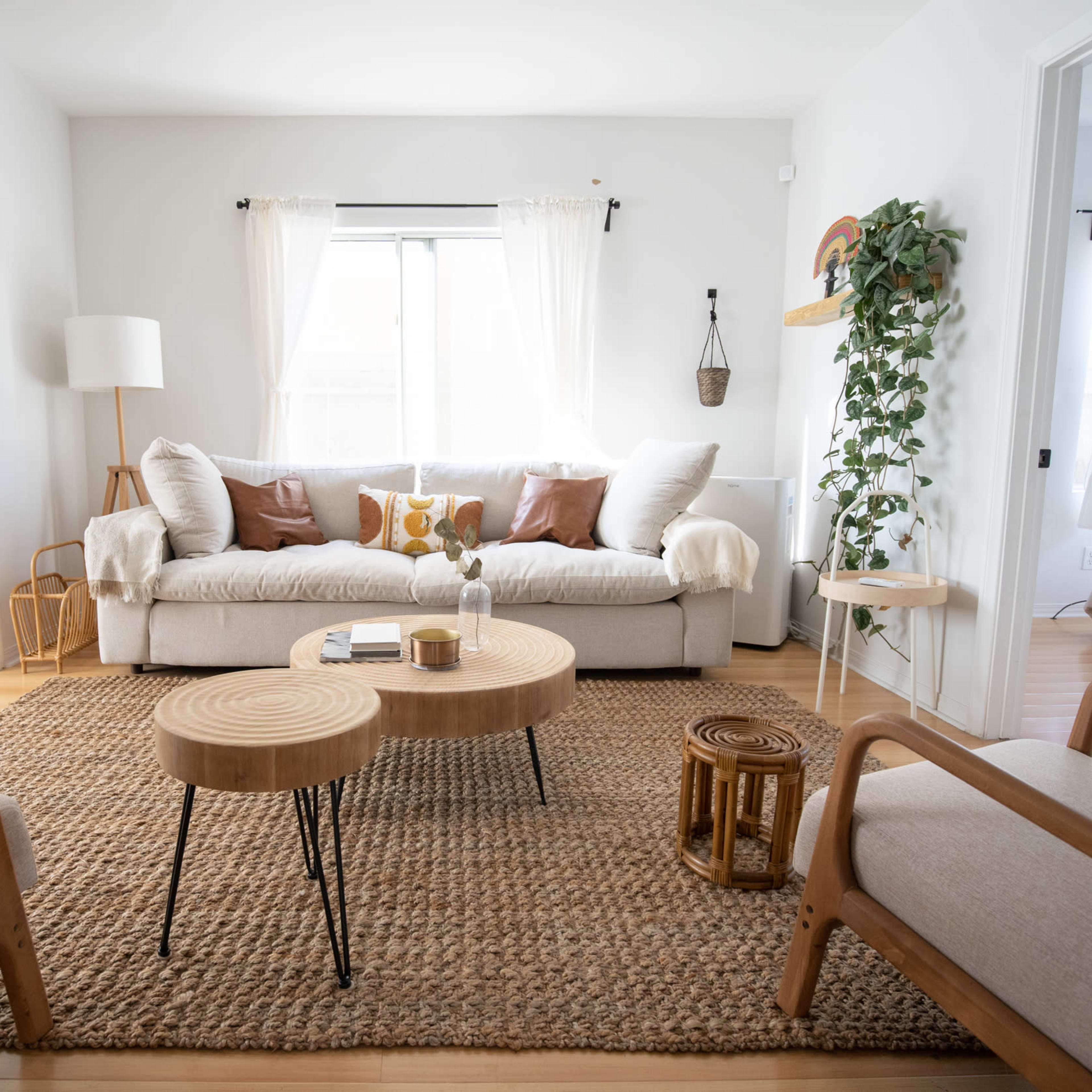 A bright living room featuring a beige couch with decorative pillows, two wooden chairs, a woven rug, and a pair of coffee tables.