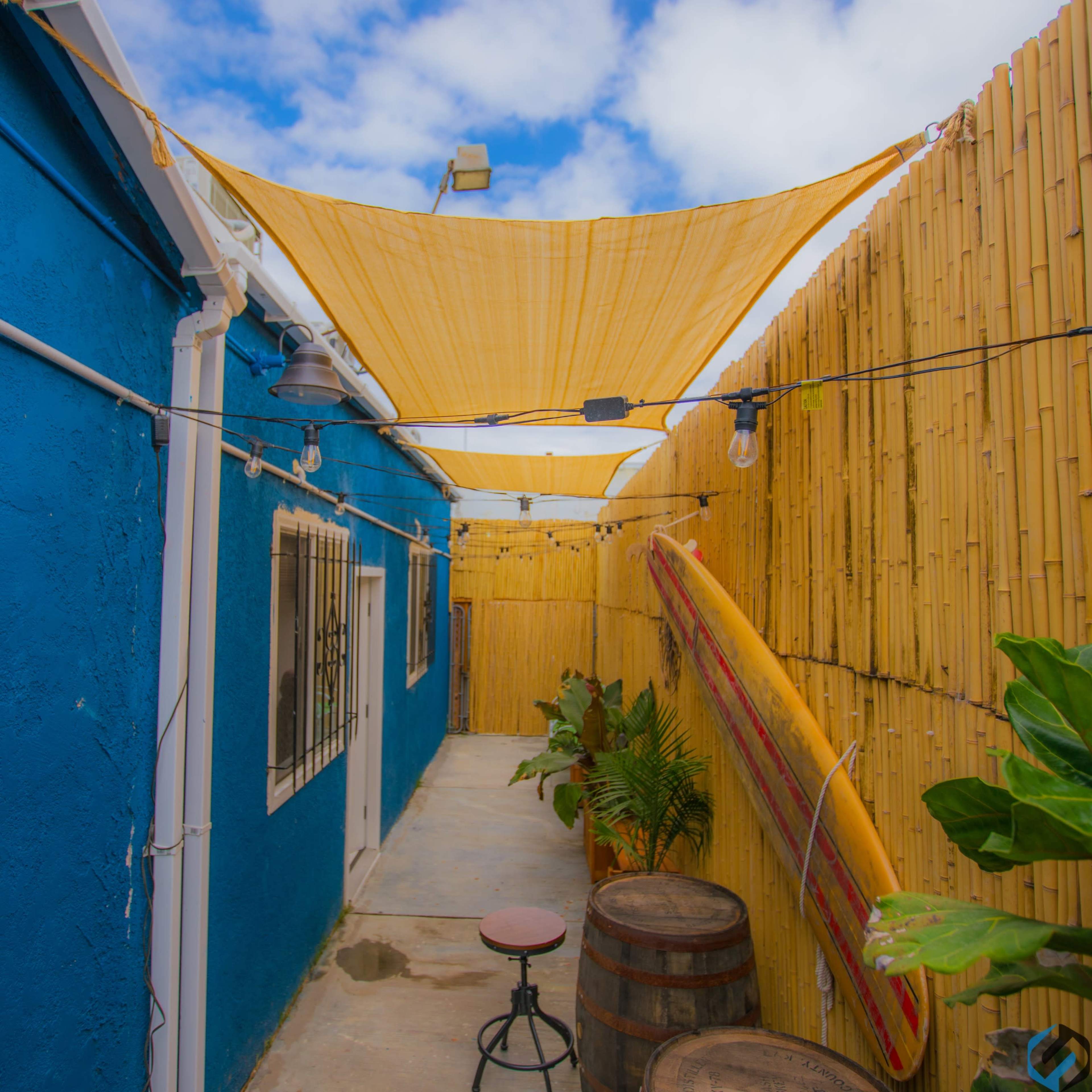 A narrow path lined with a blue wall, bamboo fencing, and hanging lights is shaded by a yellow canopy.
