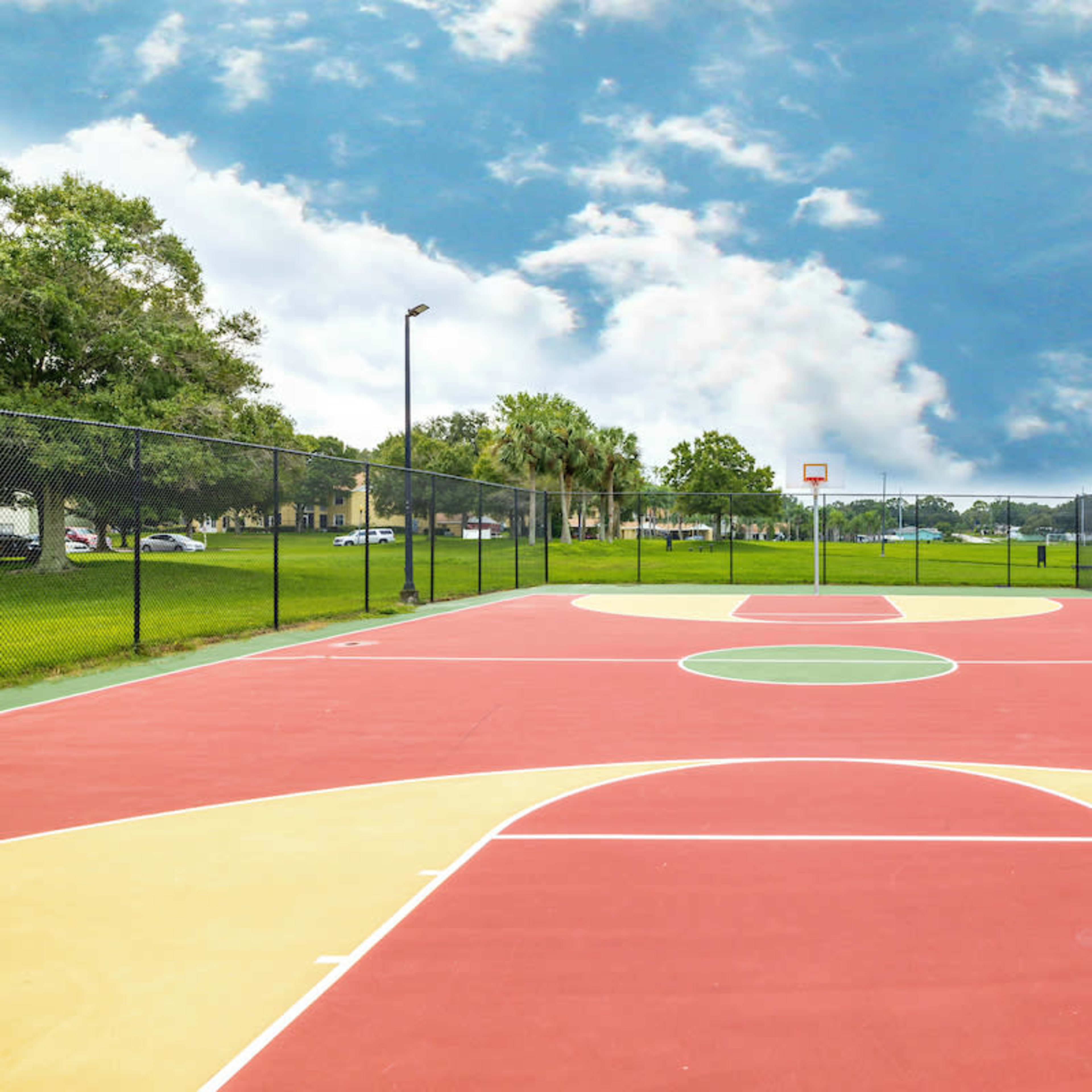 A vacant outdoor basketball court surrounded by grass and trees under a cloudy sky.