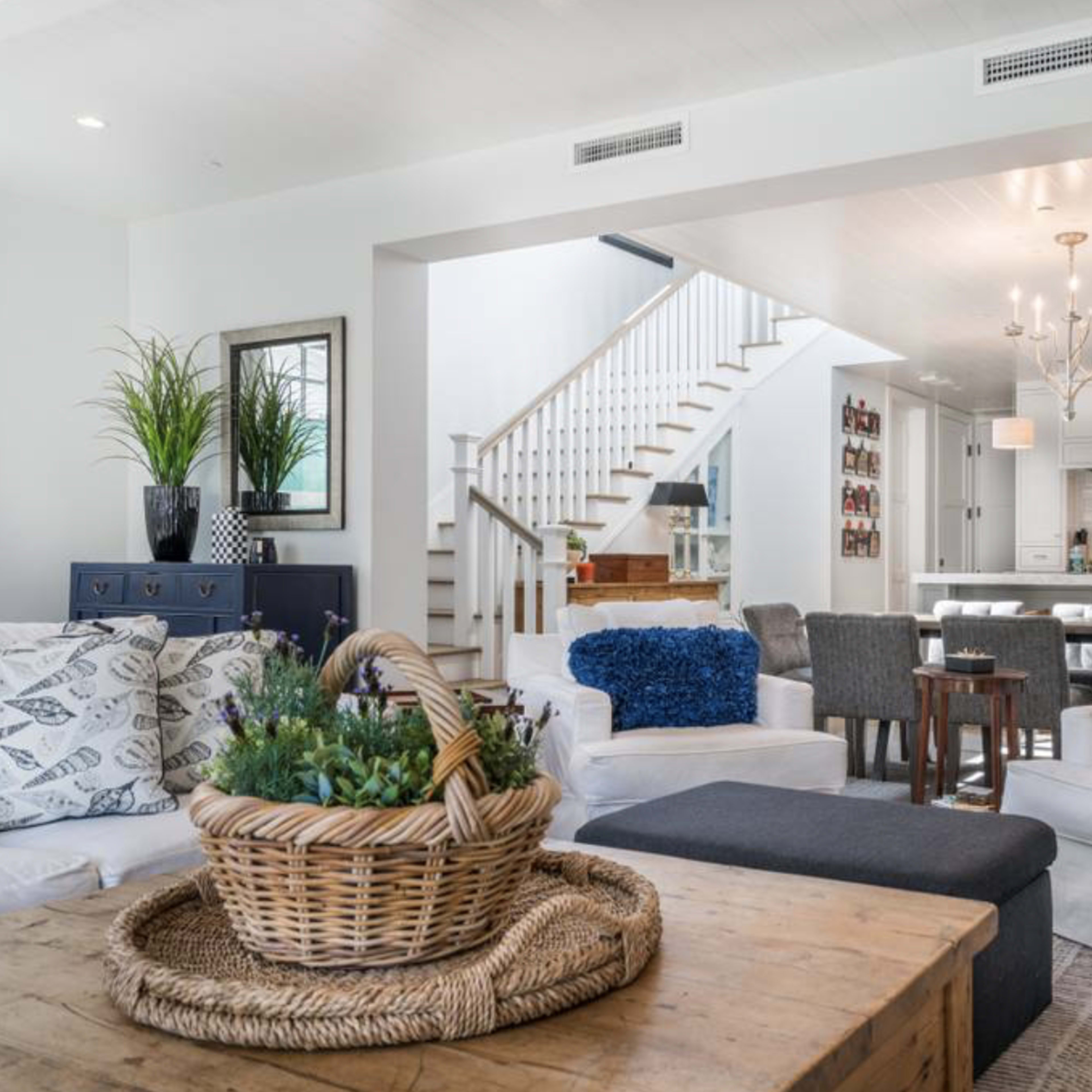 A bright living room features white sofas adorned with blue cushions, a wooden coffee table, and a staircase leading to a kitchen area in the background.