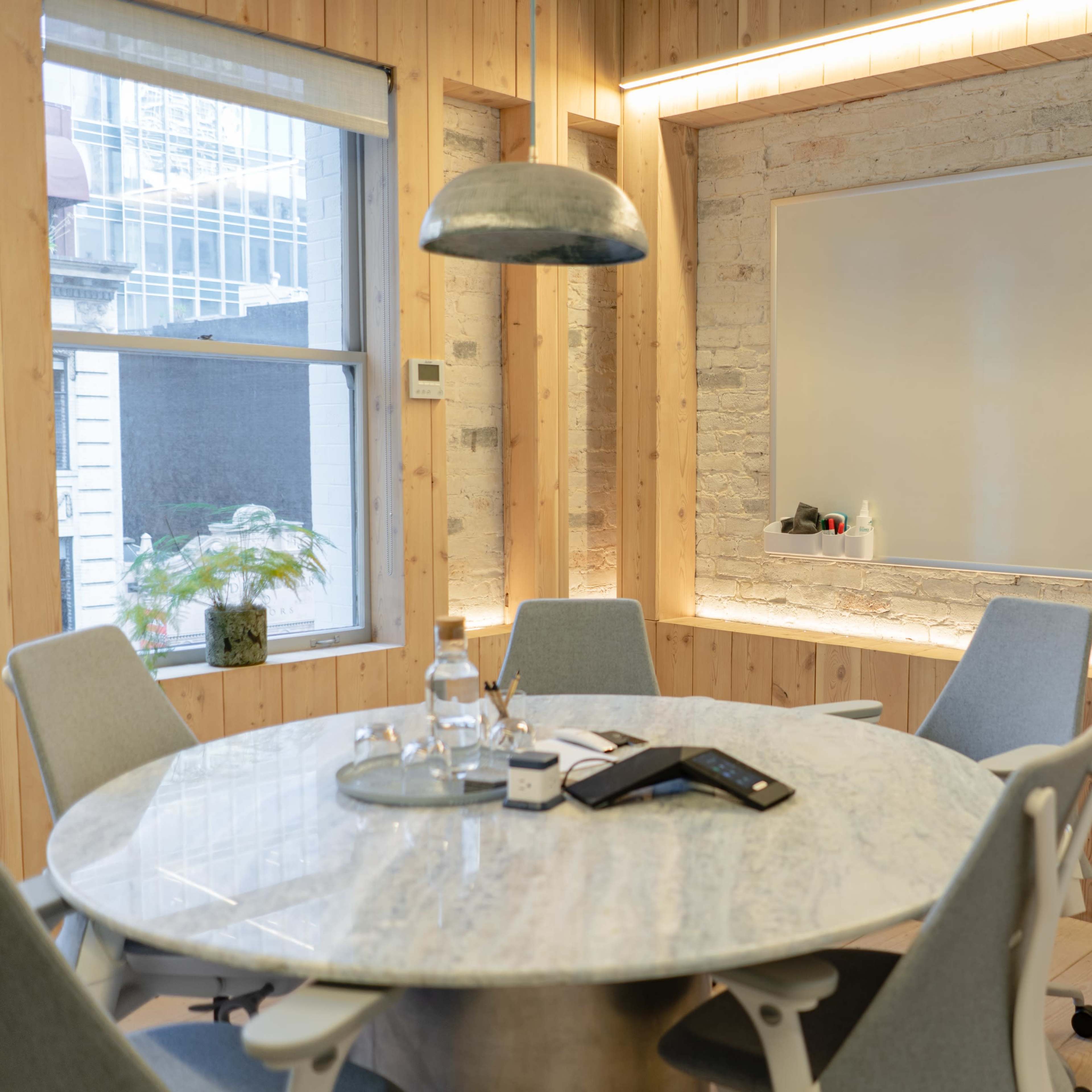 A modern conference room features a round marble table surrounded by gray chairs, with a whiteboard and large windows in the background.