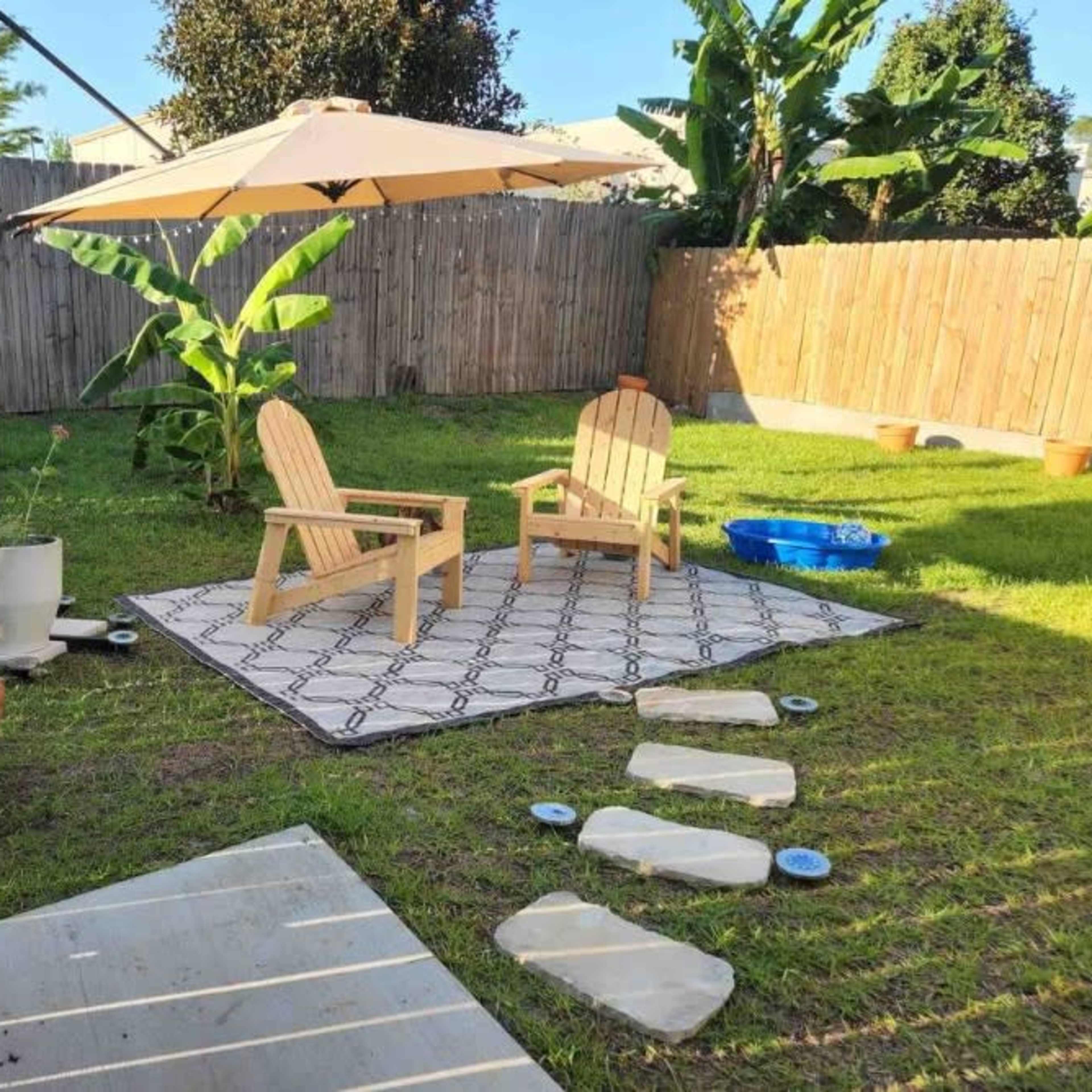 A small backyard features two wooden chairs under an umbrella, surrounded by potted plants, a blue water container, and a grass area with stepping stones.