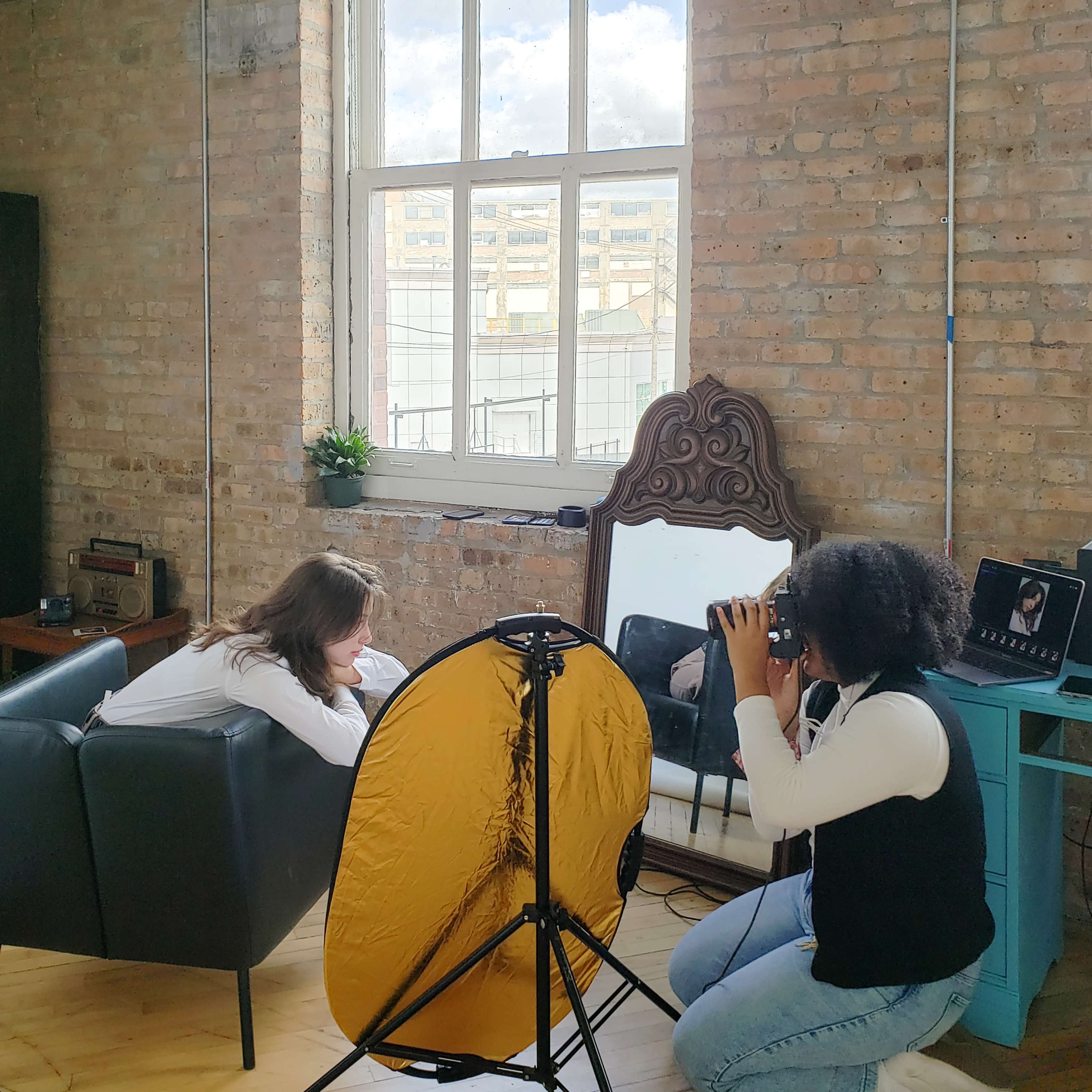 A photographer captures an image of a model sitting on a sofa in a studio setting with large windows and a backdrop.