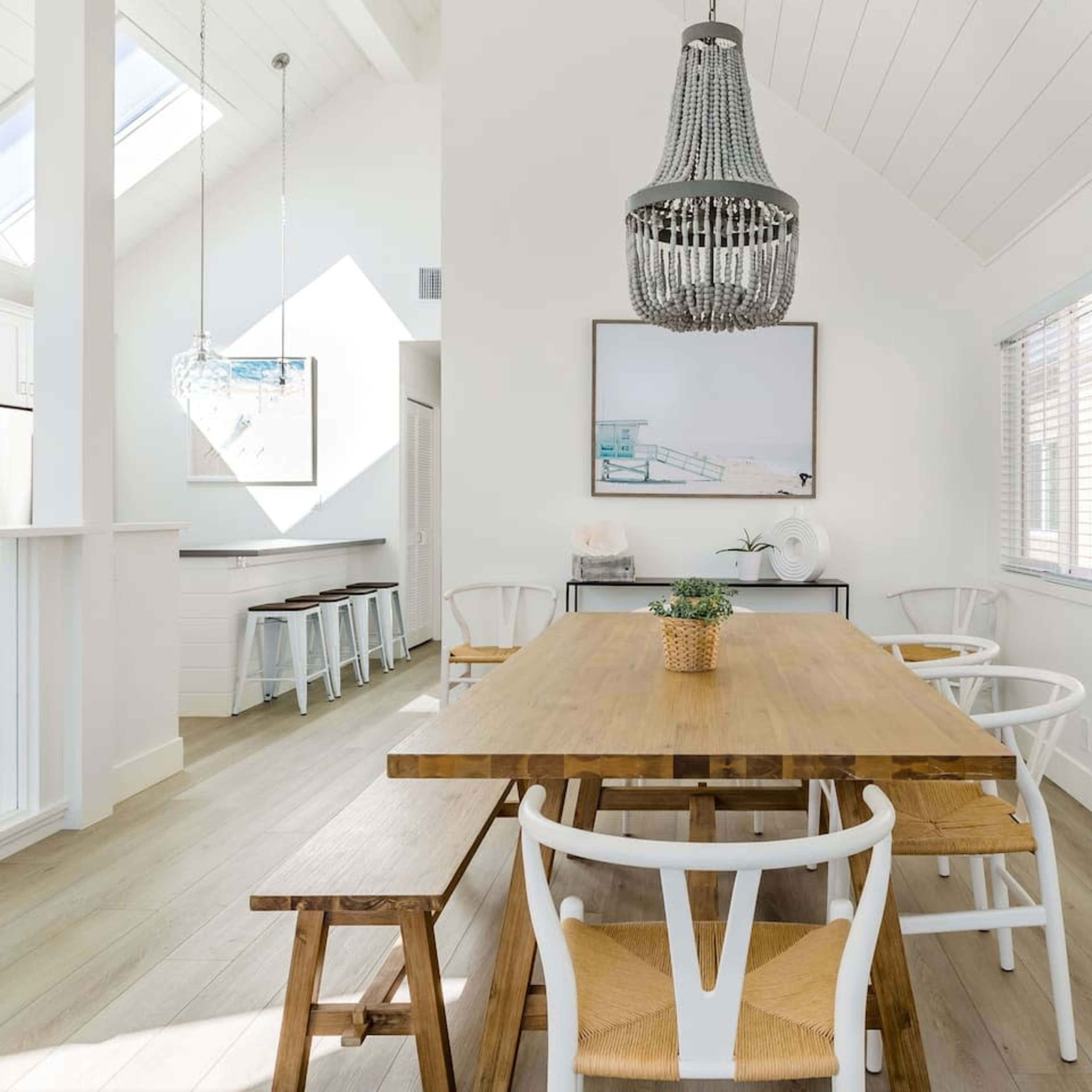 A wooden dining table with white chairs is set in a bright room featuring large windows and a chandelier.