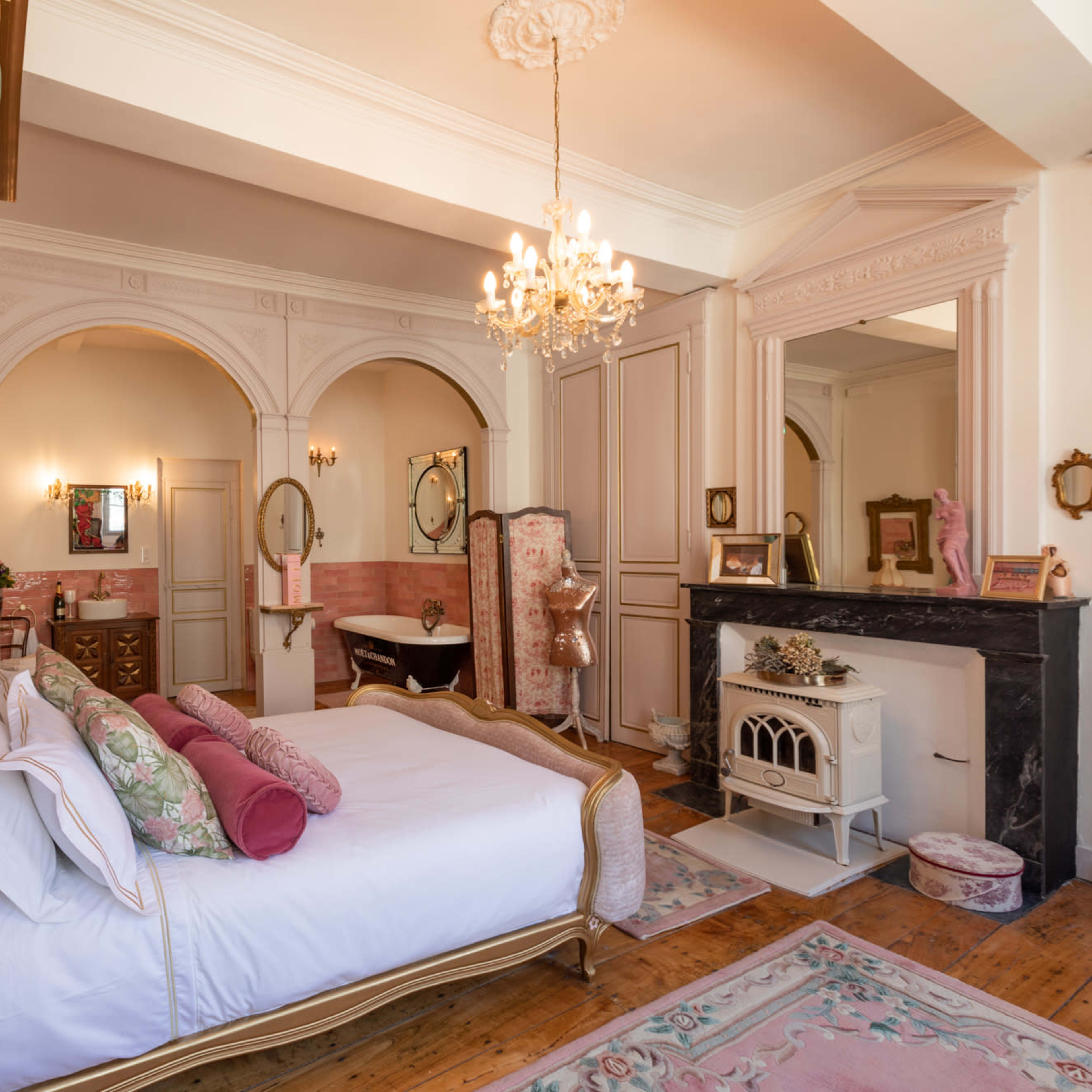 A elegantly decorated bedroom featuring a large bed, vintage-style mirrors, a chandelier, and a decorative fireplace.
