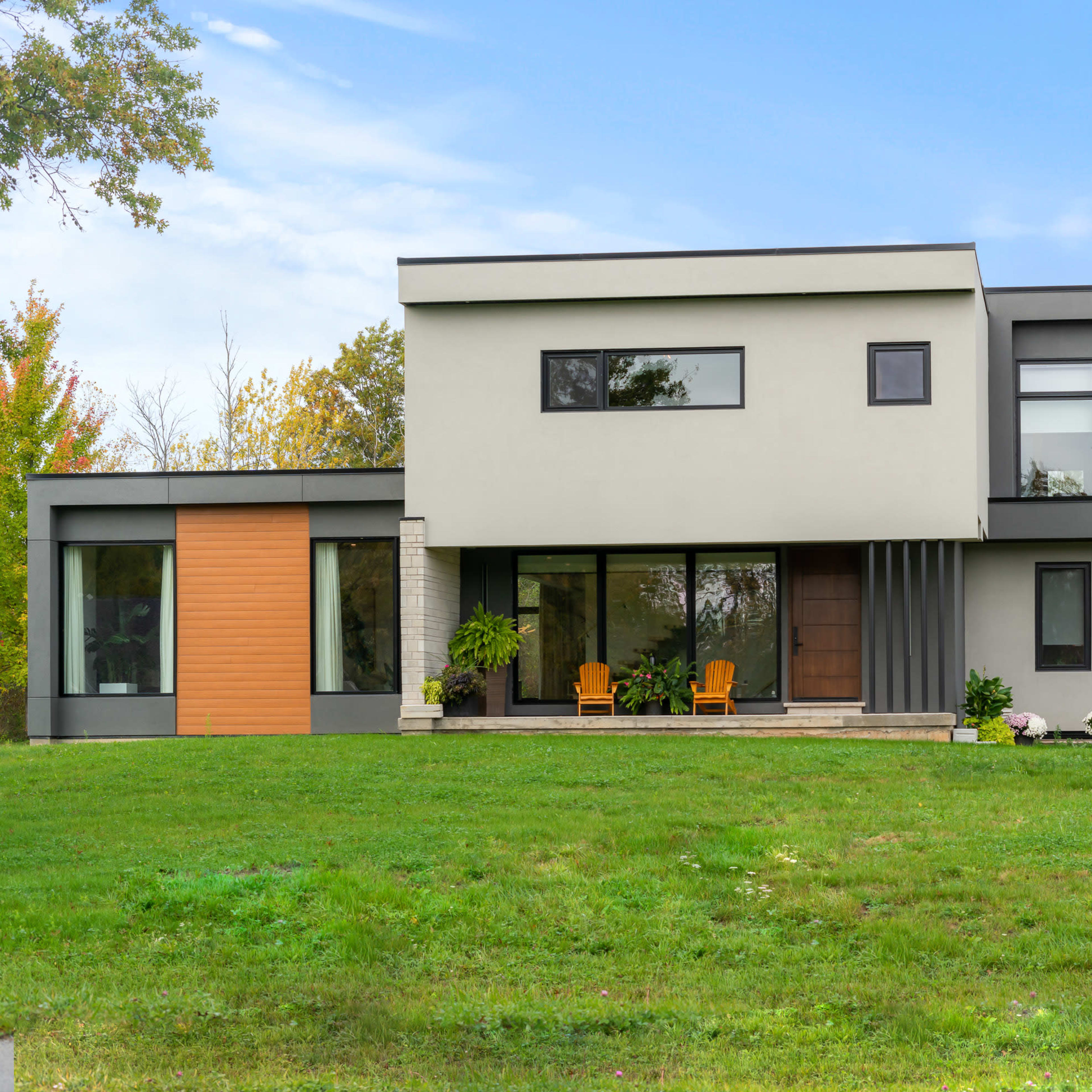 A modern two-story house features a mix of gray and wooden panel exteriors, large windows, and a front porch with two chairs.