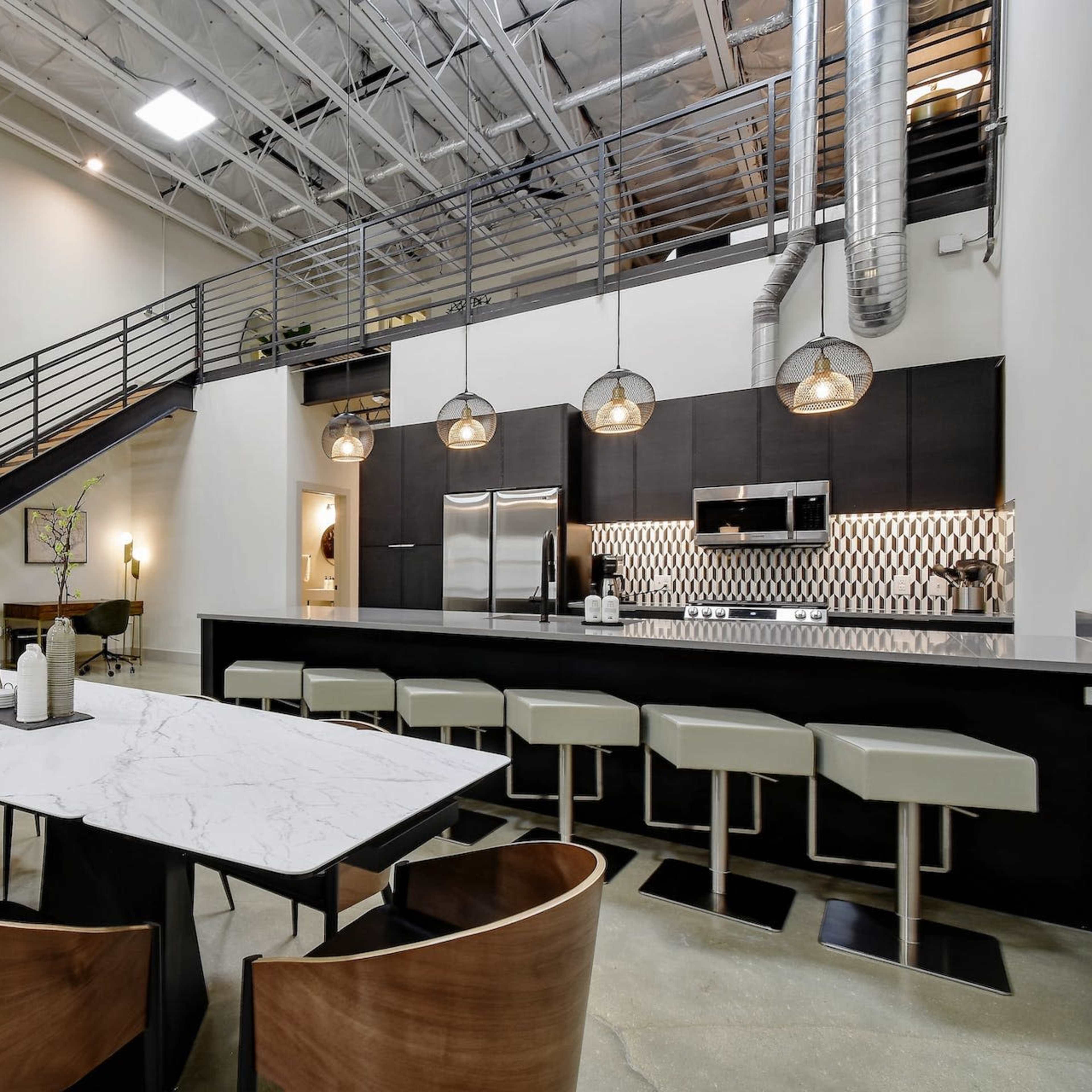 A modern open-concept kitchen features a marble dining table surrounded by wooden chairs, with a staircase in the background and pendant lights above the counter.