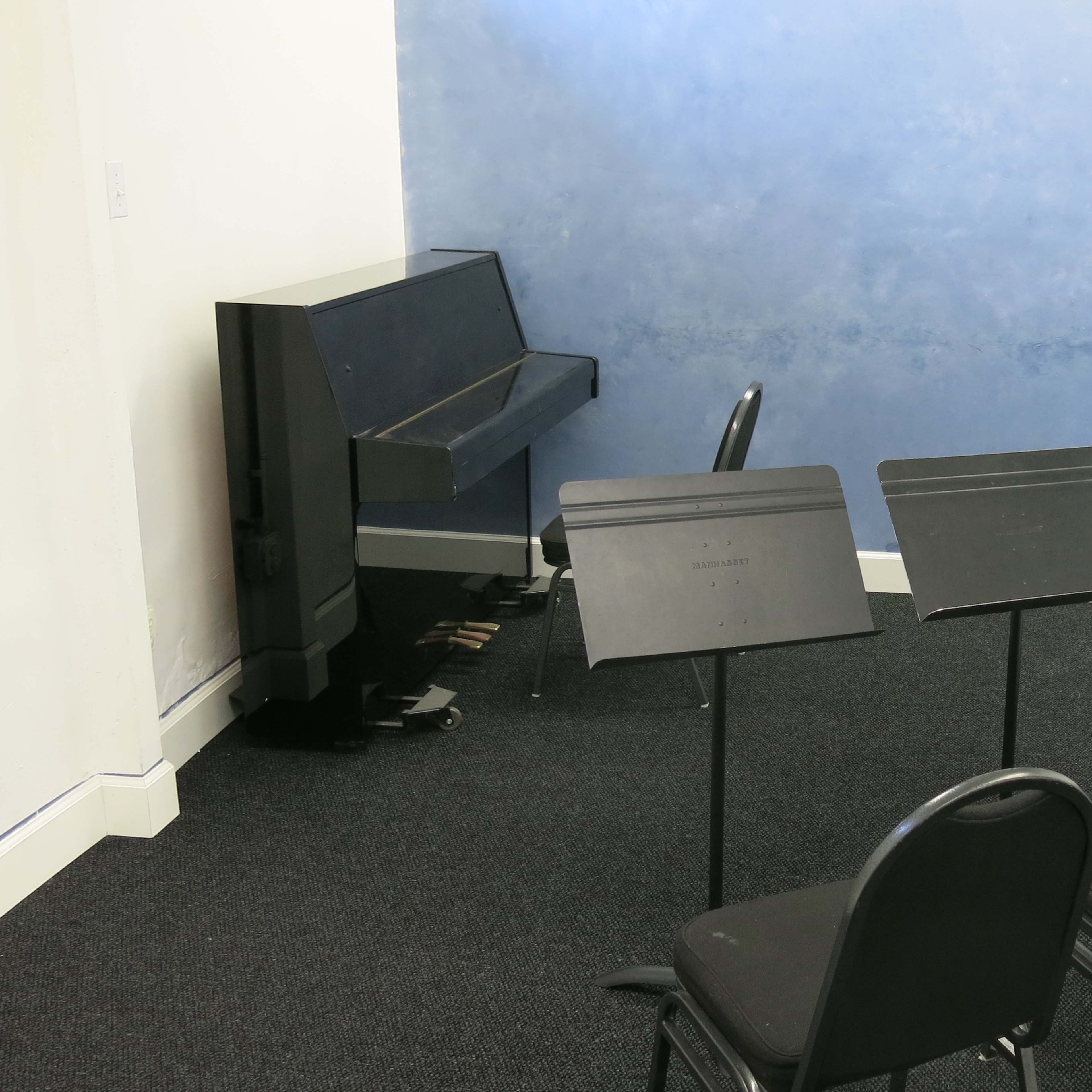 A small room with a black piano, three music stands, and a chair against a blue wall.